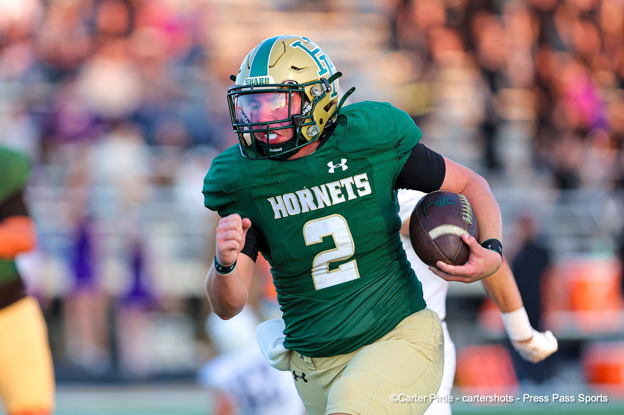 Football player in a green Hornets uniform runs with the ball during a game, crowd blurred in the background.
