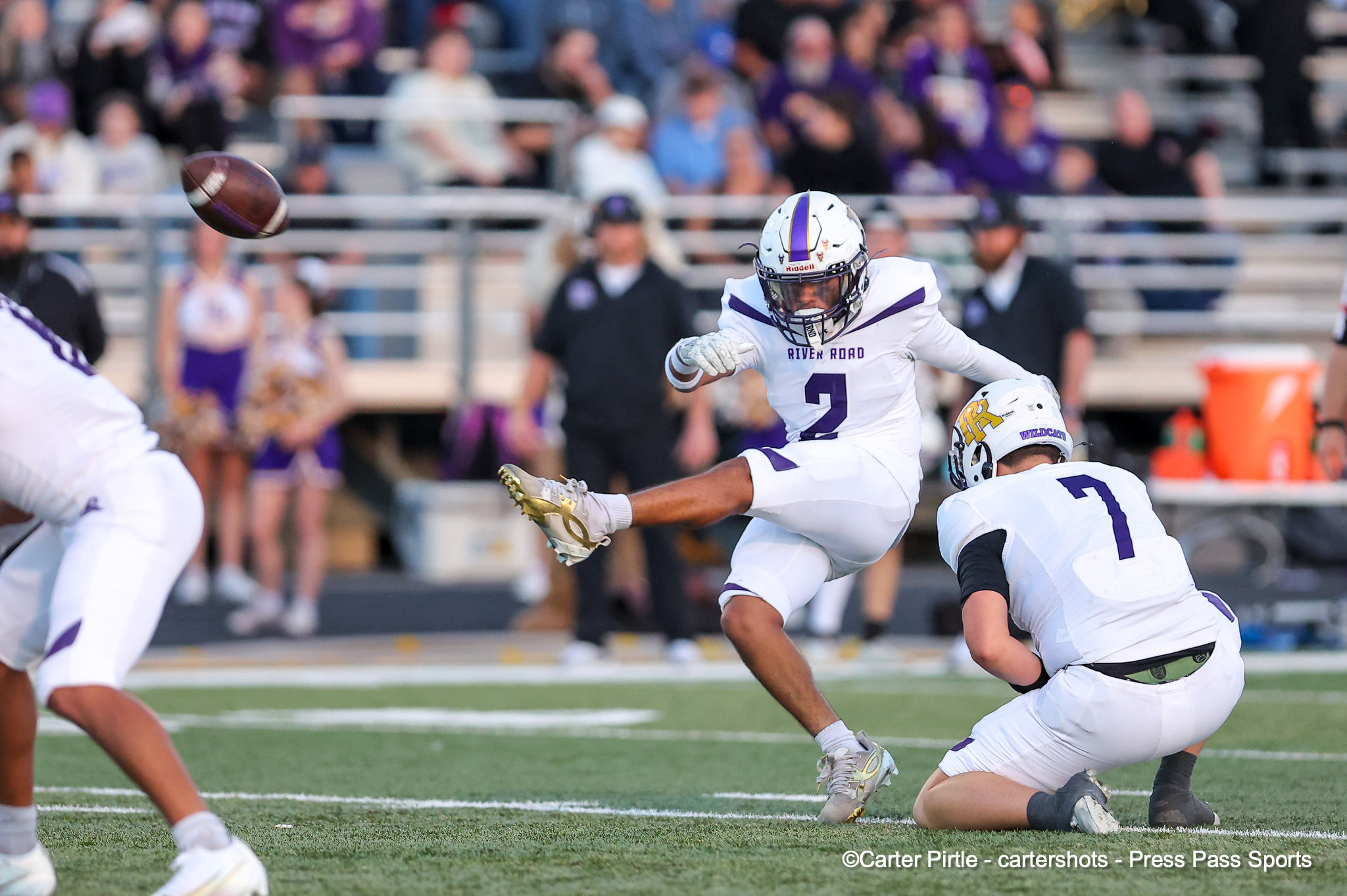 Football players in white and purple chase an airborne ball during a game, with a crowd in the stands behind them.