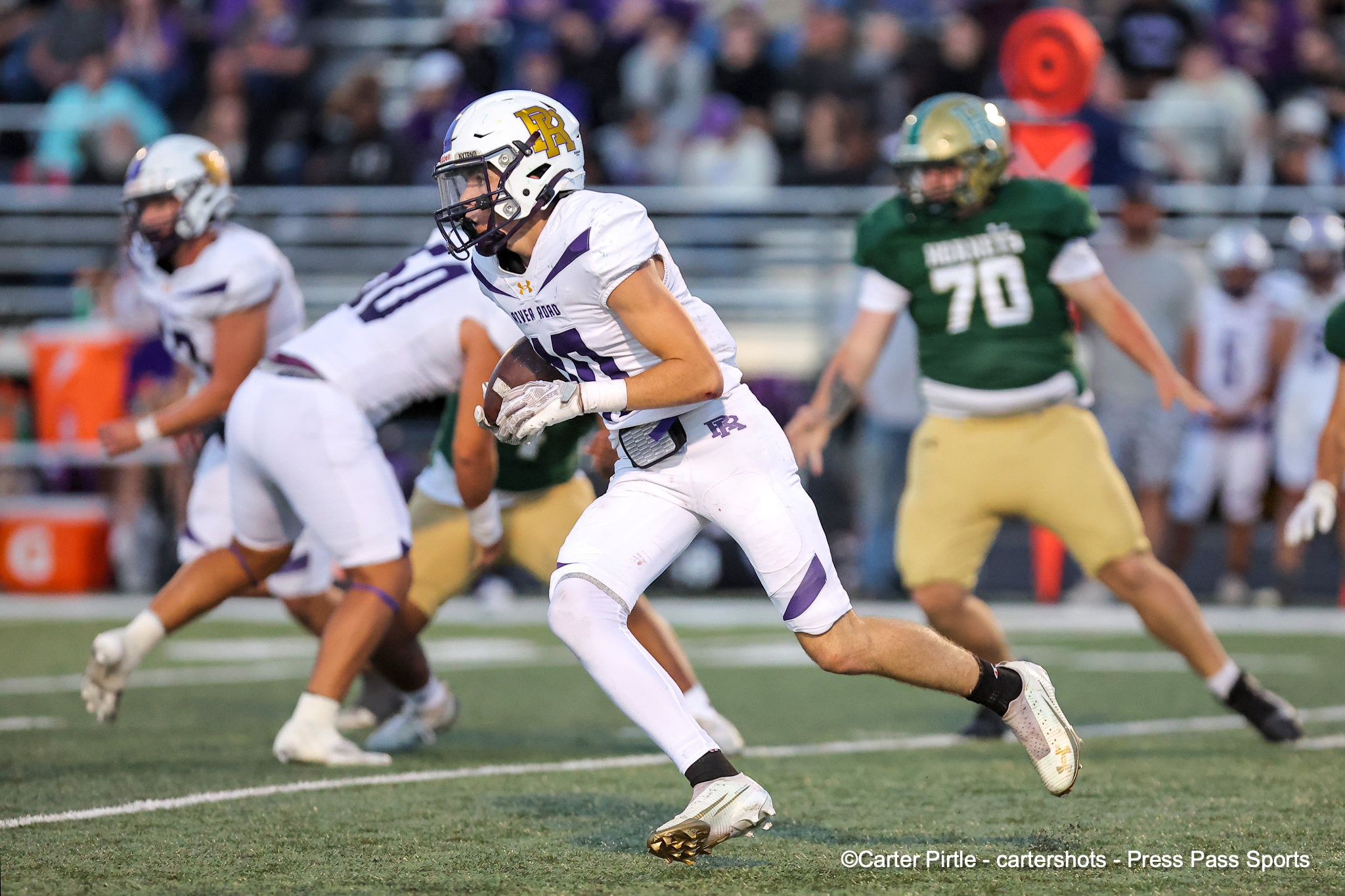 Football player in a white uniform runs with the ball during a game, pursued by defenders in green.