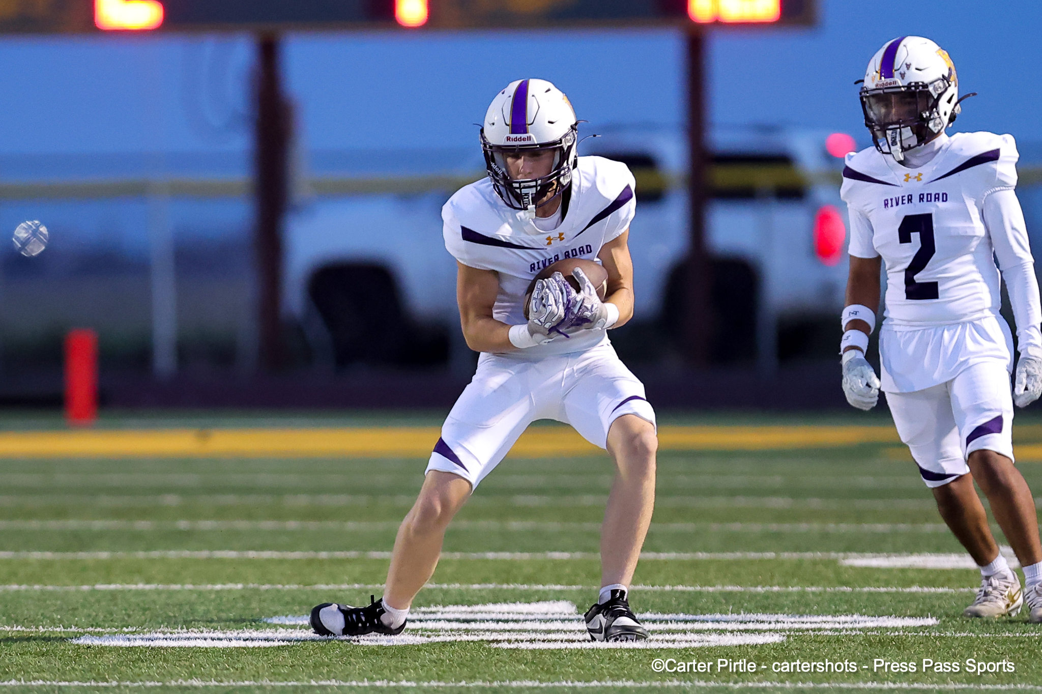 Two football players in white and purple uniforms on a field; one carries the ball as he runs, another teammate nearby.