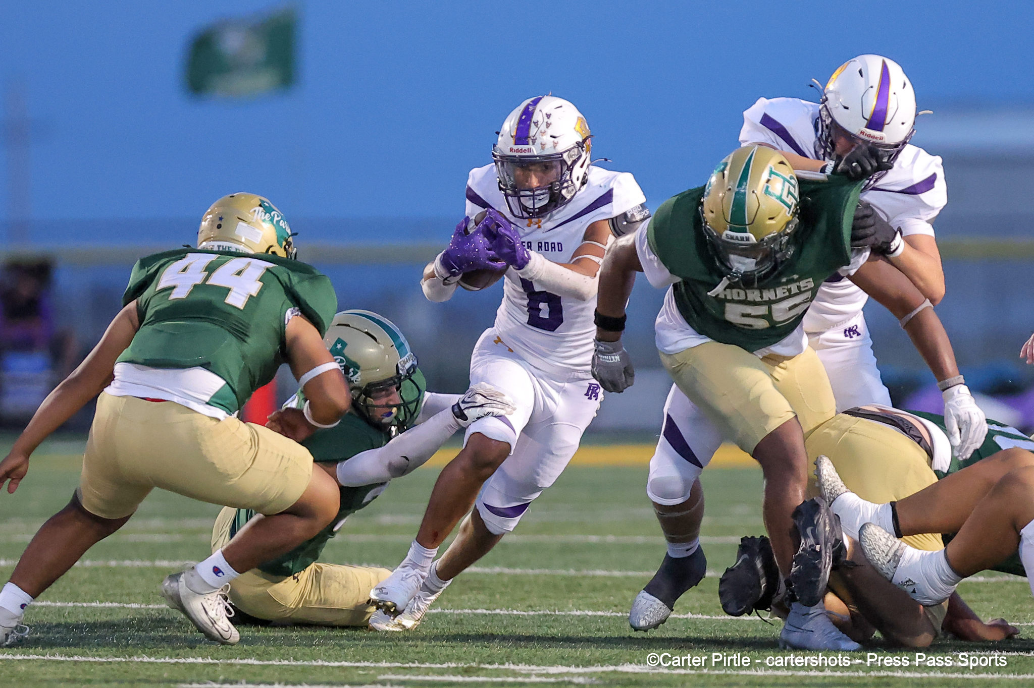 A group of football players in green and gold collide with players in white and purple during a play on a grassy field.