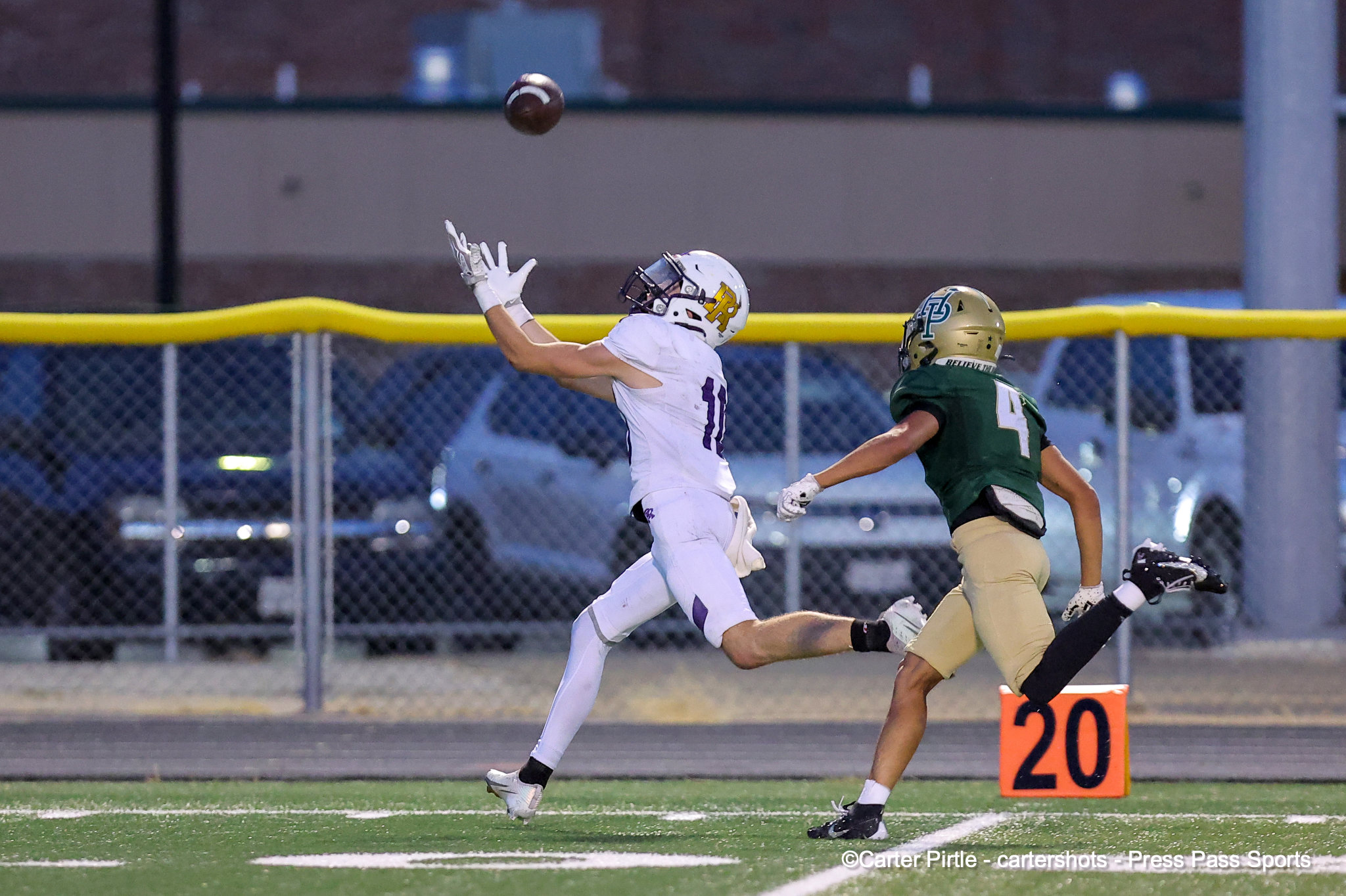 Two football players leap for a high ball near the end zone as a defender in white reaches up while an opponent in green and gold closes in.