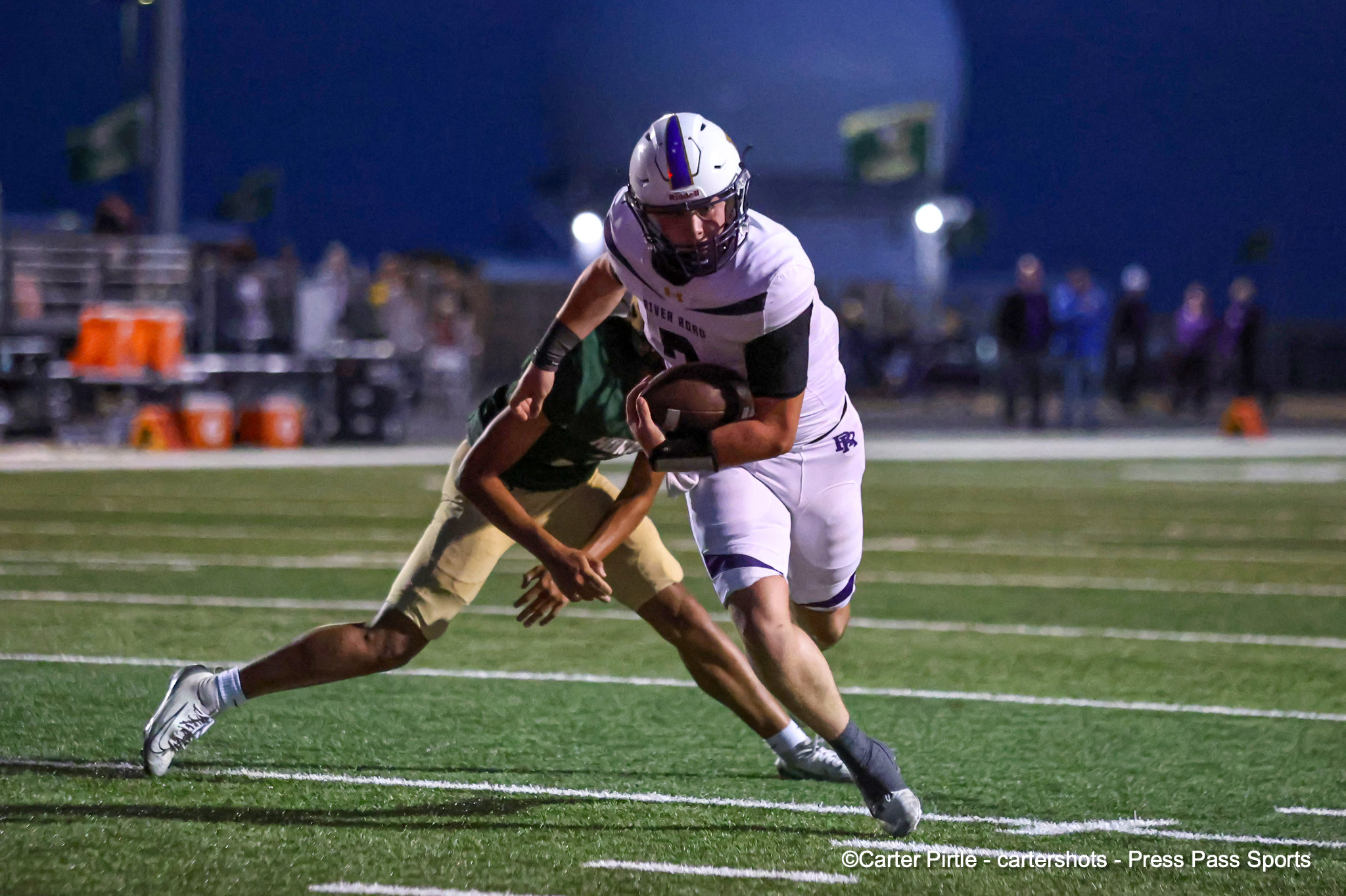 A football player in white uniform being tackled from behind on a turf field at night, holding the ball tightly.