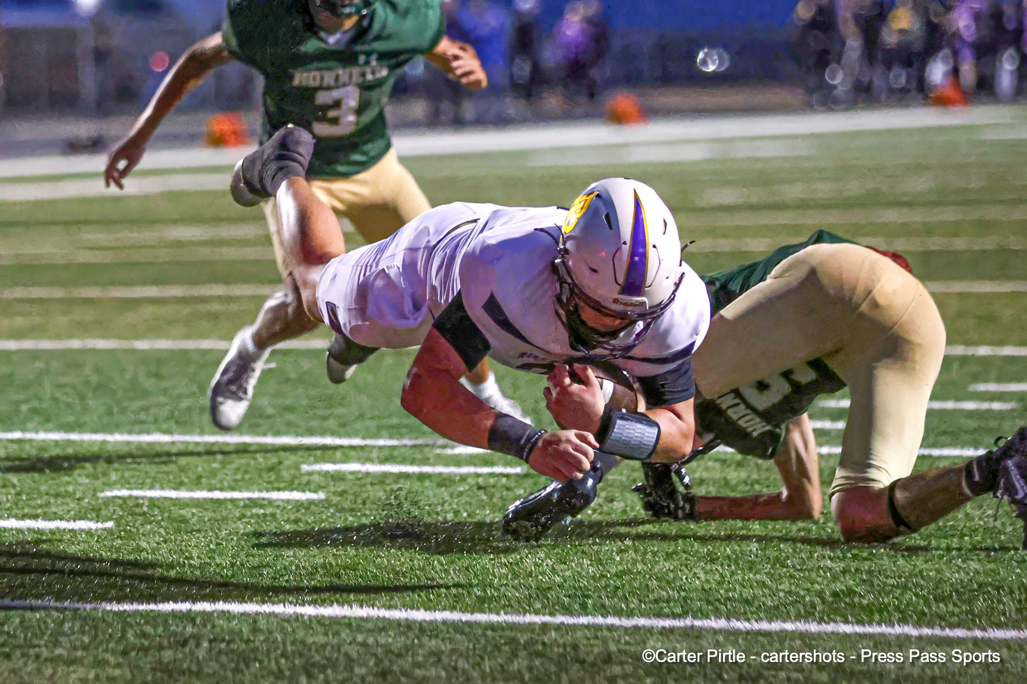 Two football players collide during a tackle on a green turf field at night, with yard lines visible in the foreground and spectators blurred in the background.