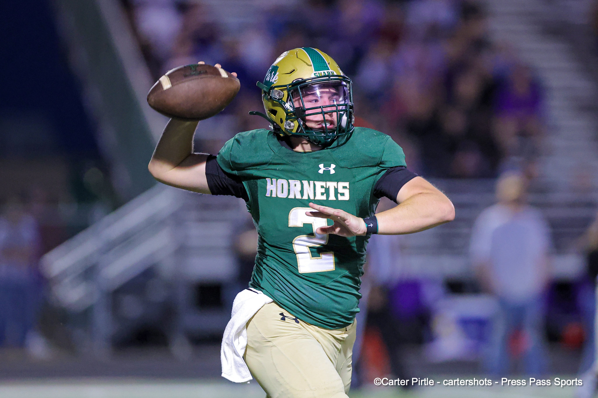 Football quarterback in a green Hornets jersey throws a pass as crowds blur in the background, helmet on and arm cocked back.