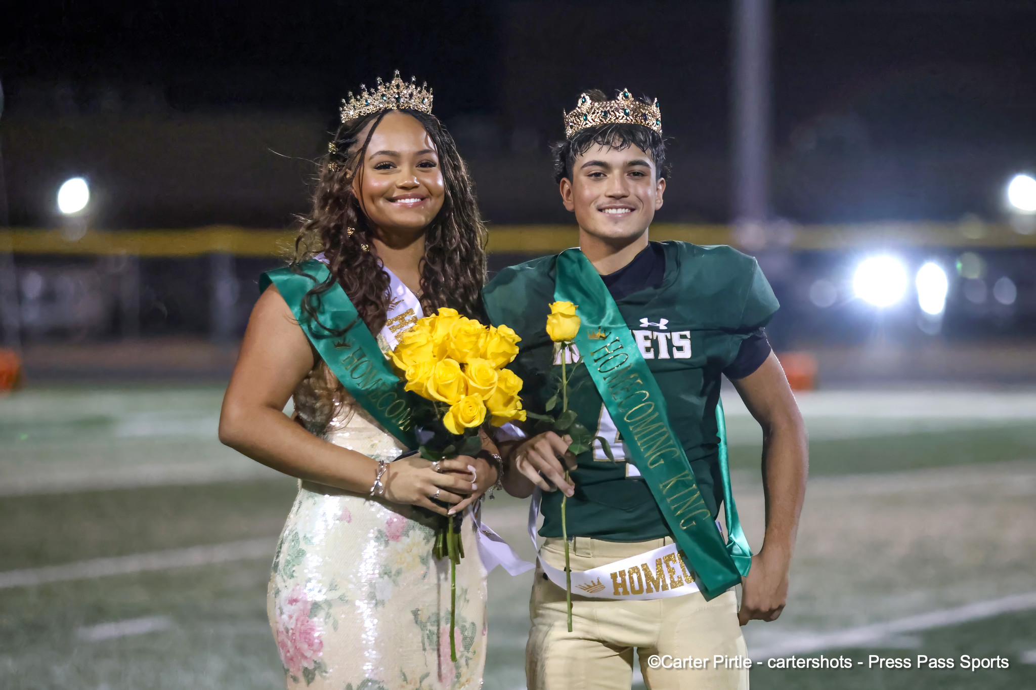 Two pageant winners wearing crowns and green sashes pose on a football field at night, holding yellow roses and smiling at the camera.