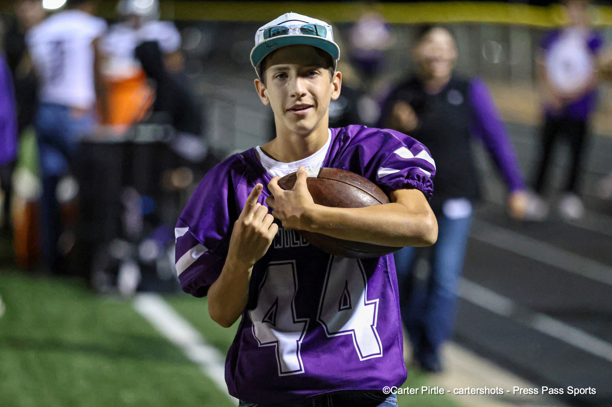 Young football player in a purple jersey with the number 44, running with a football tucked under his arm on a track or field.