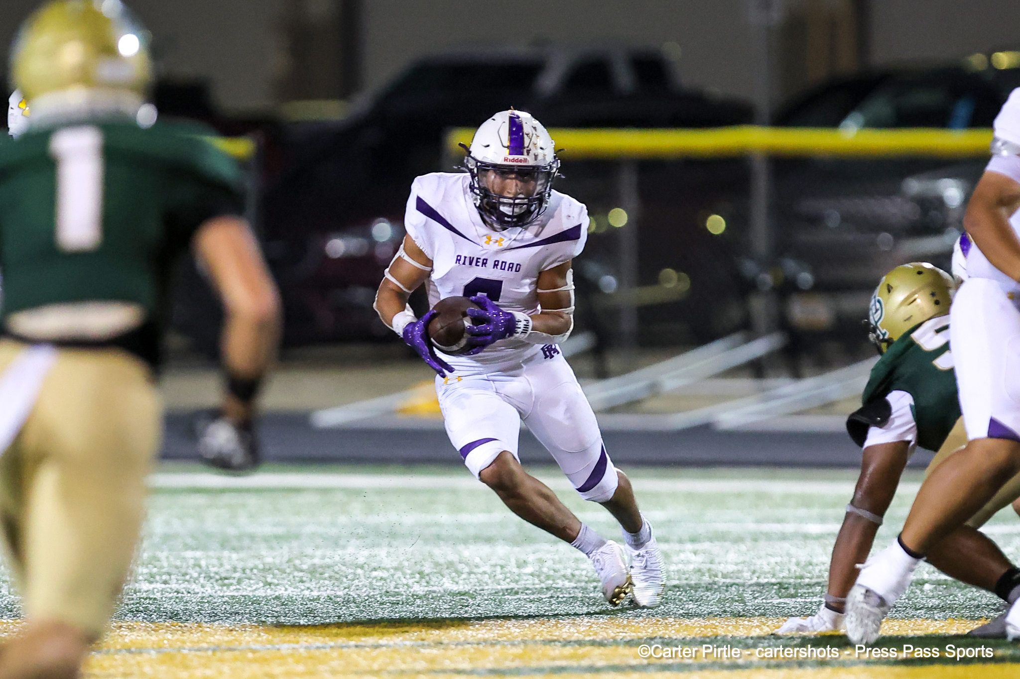 Football player in white and purple jersey with'River Road' runs with the ball, surrounded by defenders at night.