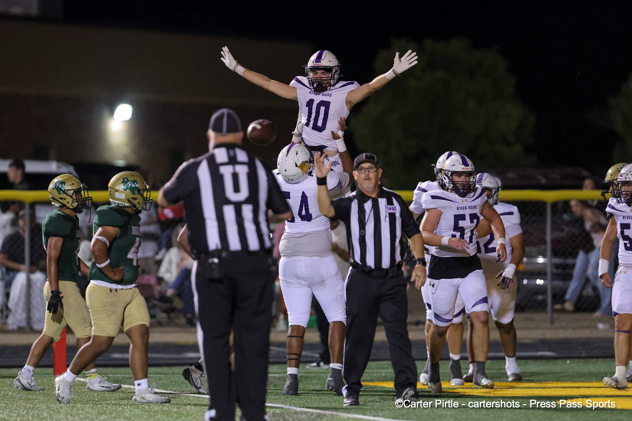 Night football celebration: a purple-and-white team lifts a teammate wearing #10 on their shoulders while officials and other players look on.