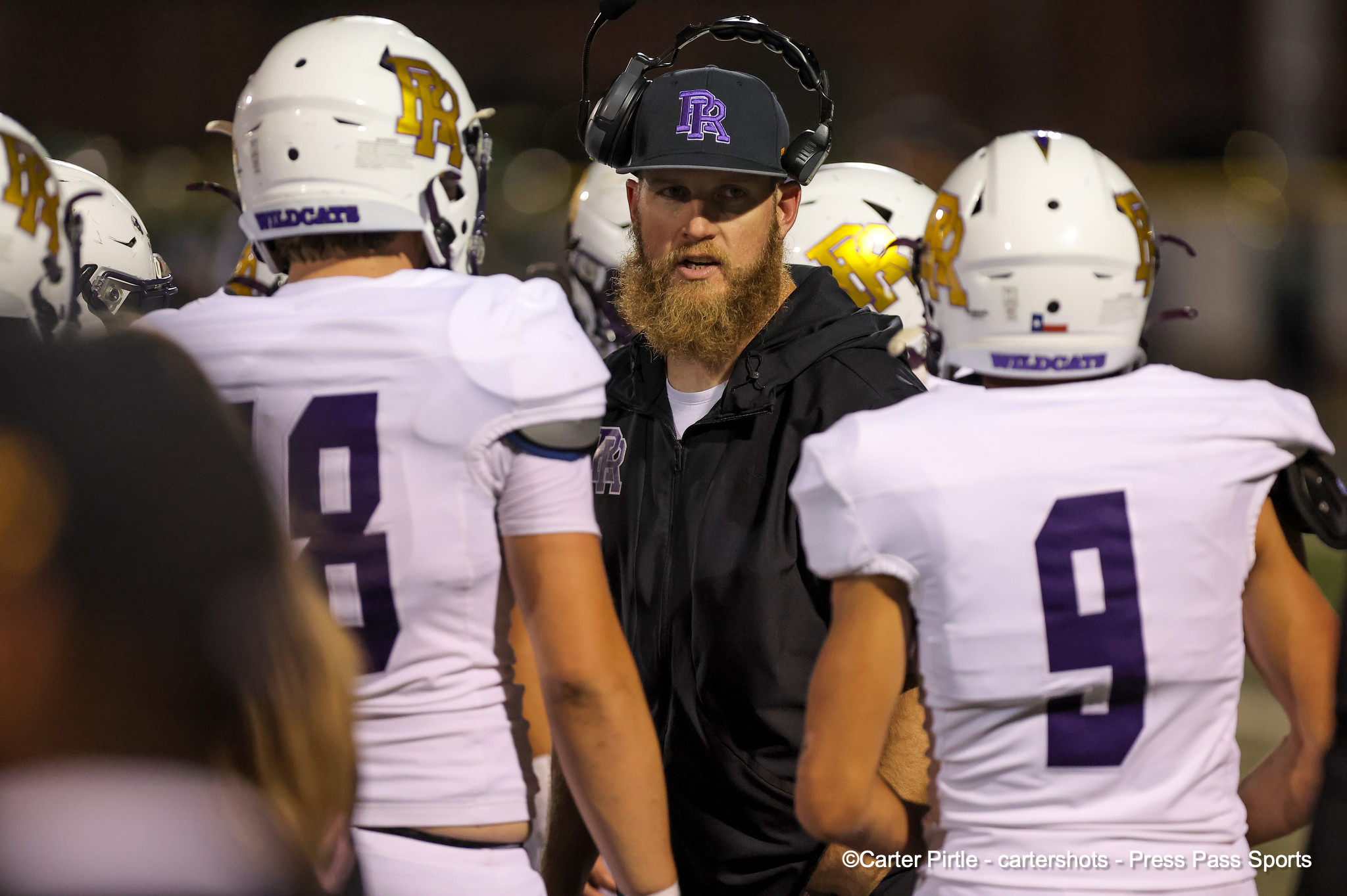 Football coach with headset and purple-logo cap addressing players on a night practice.