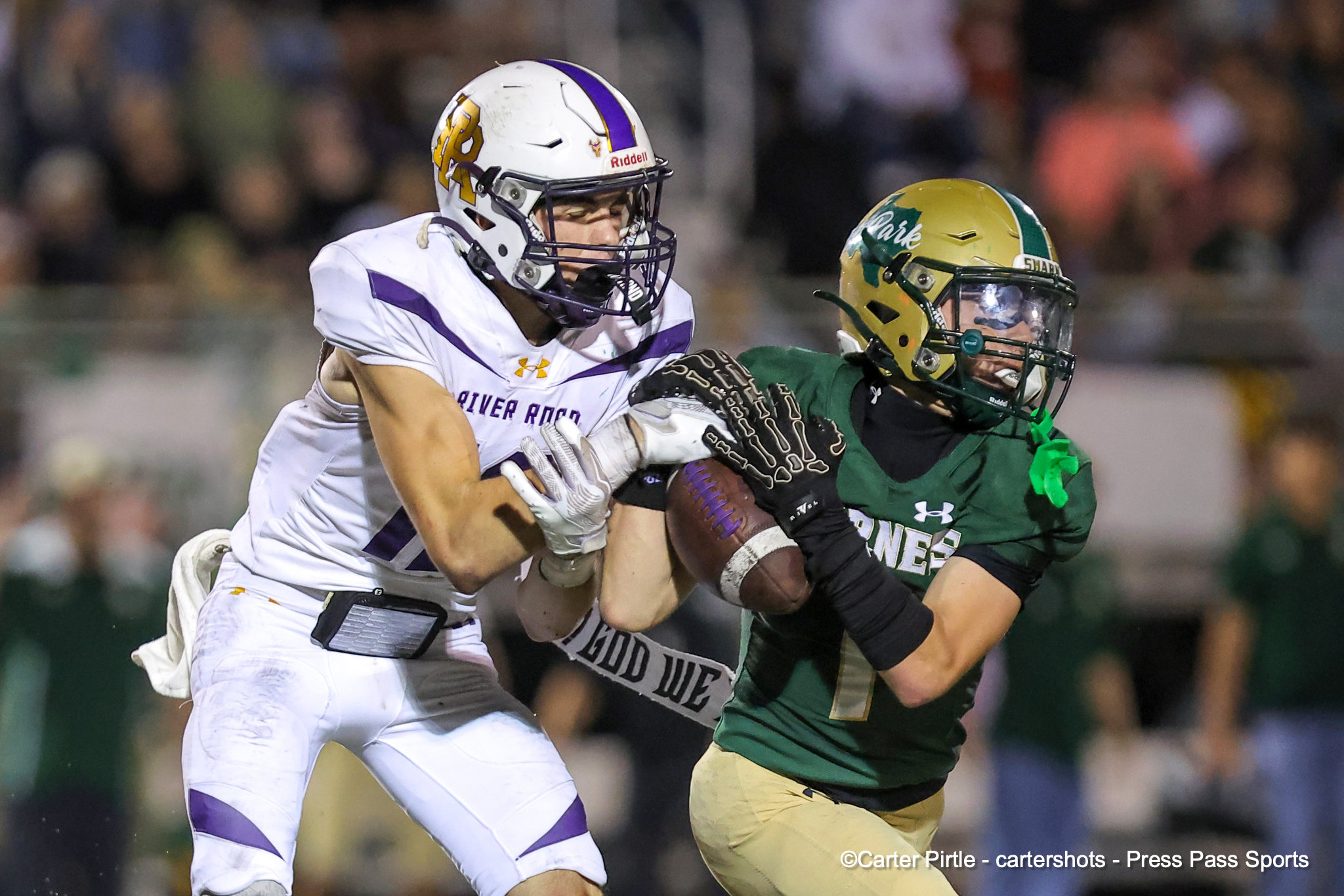 Two football players collide in a game: one in a white and purple uniform, the other in green and gold, both wearing helmets and pads beneath a blurred crowd backdrop.