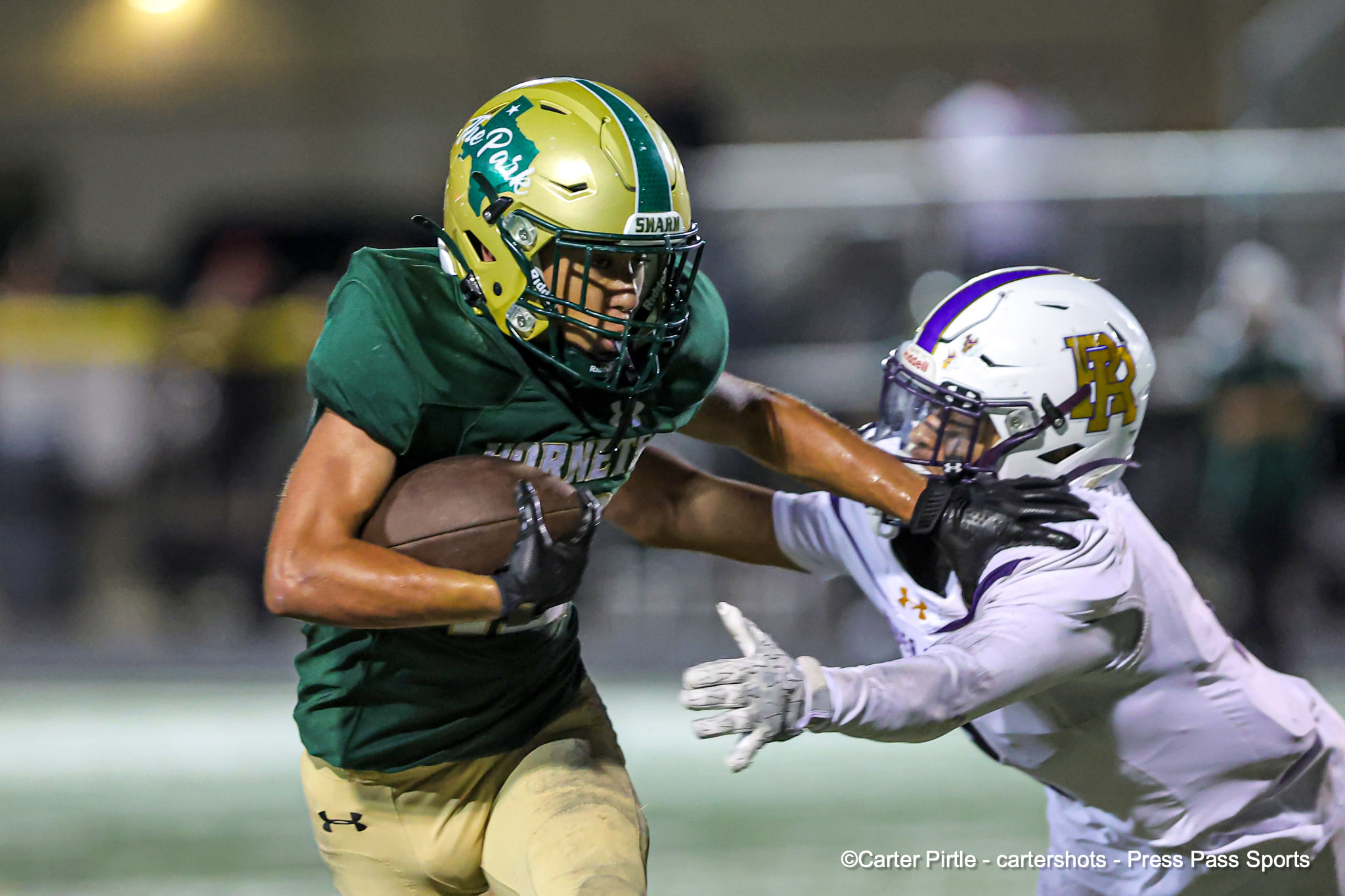 Two football players clash on a night game: one in green uniform with a gold helmet driving the ball, the other in white with purple accents reaching to tackle.