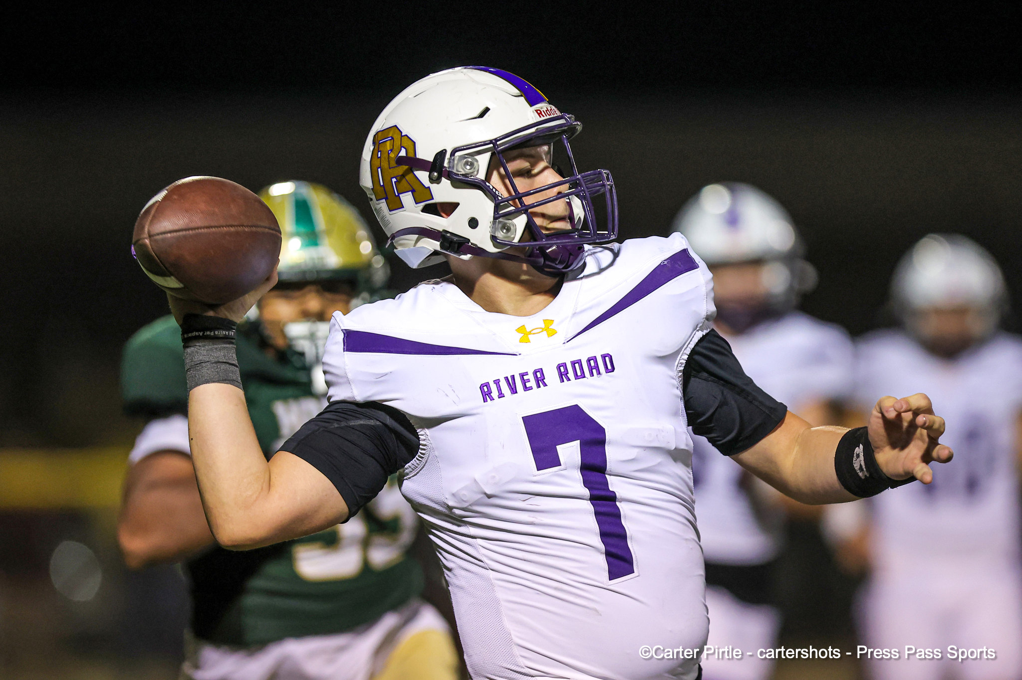 Quarterback in a white and purple River Road uniform throws a pass during a night game, helmeted and in motion.