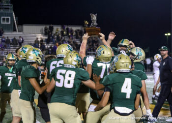 Green-clad football team lifts trophy on the field in celebration at night, stands full of fans in the background.