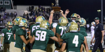 Green-clad football team lifts trophy on the field in celebration at night, stands full of fans in the background.