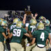 Green-clad football team lifts trophy on the field in celebration at night, stands full of fans in the background.