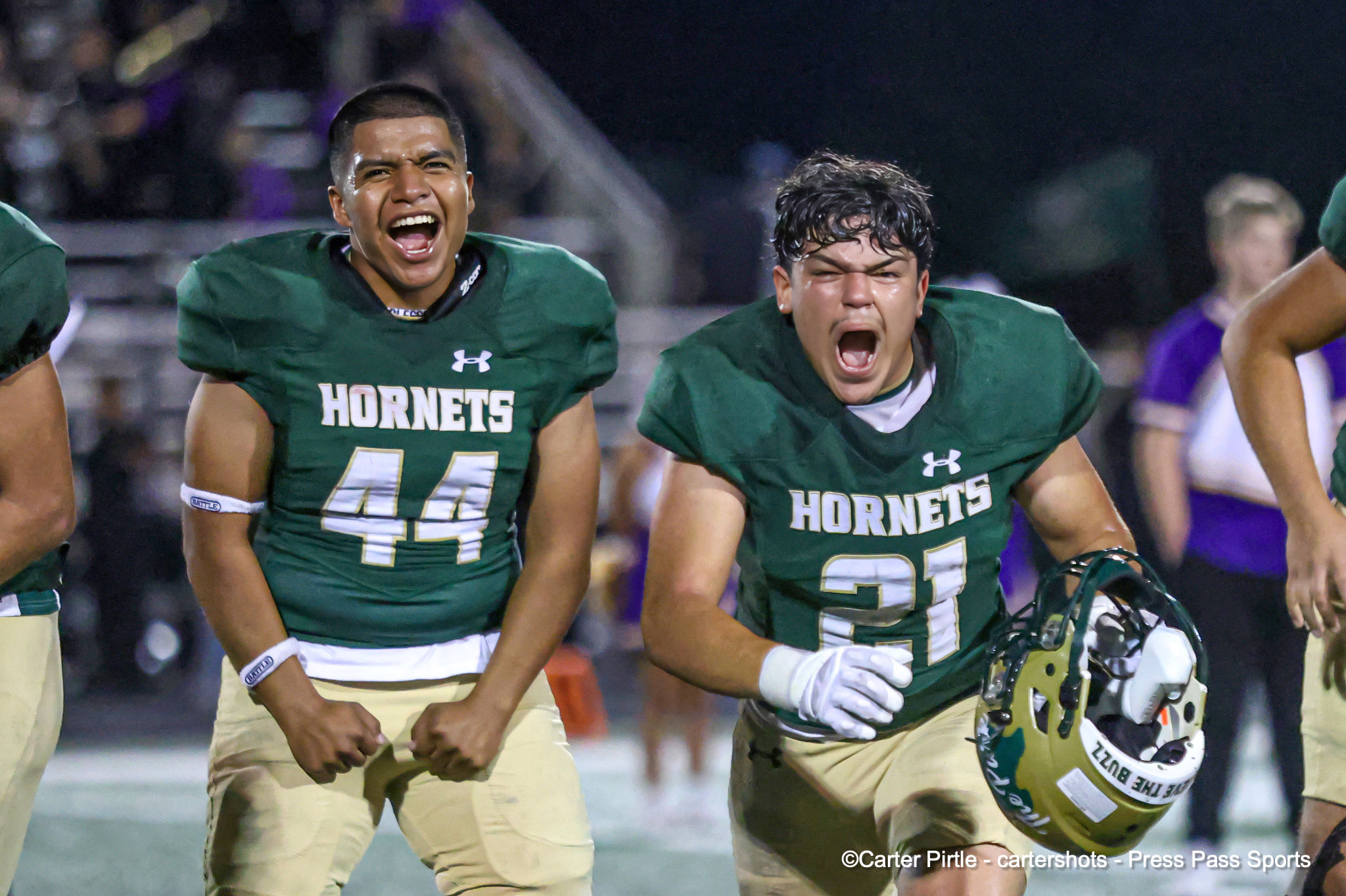 Two green Hornets football players celebrate on the field, mouths open in excitement, night game under lights.
