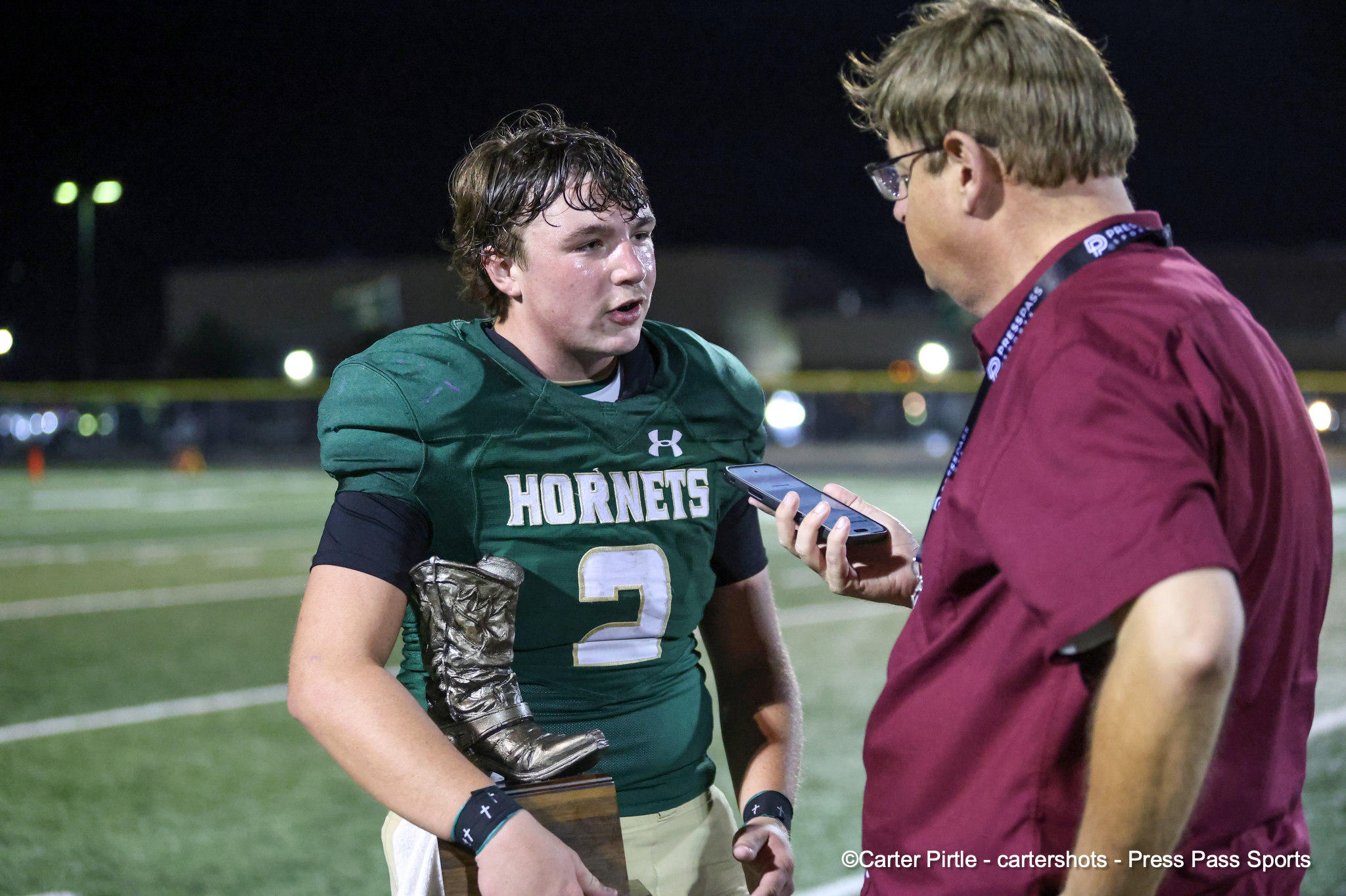 Football player in a green Hornets jersey being interviewed by a reporter holding a smartphone on a night field