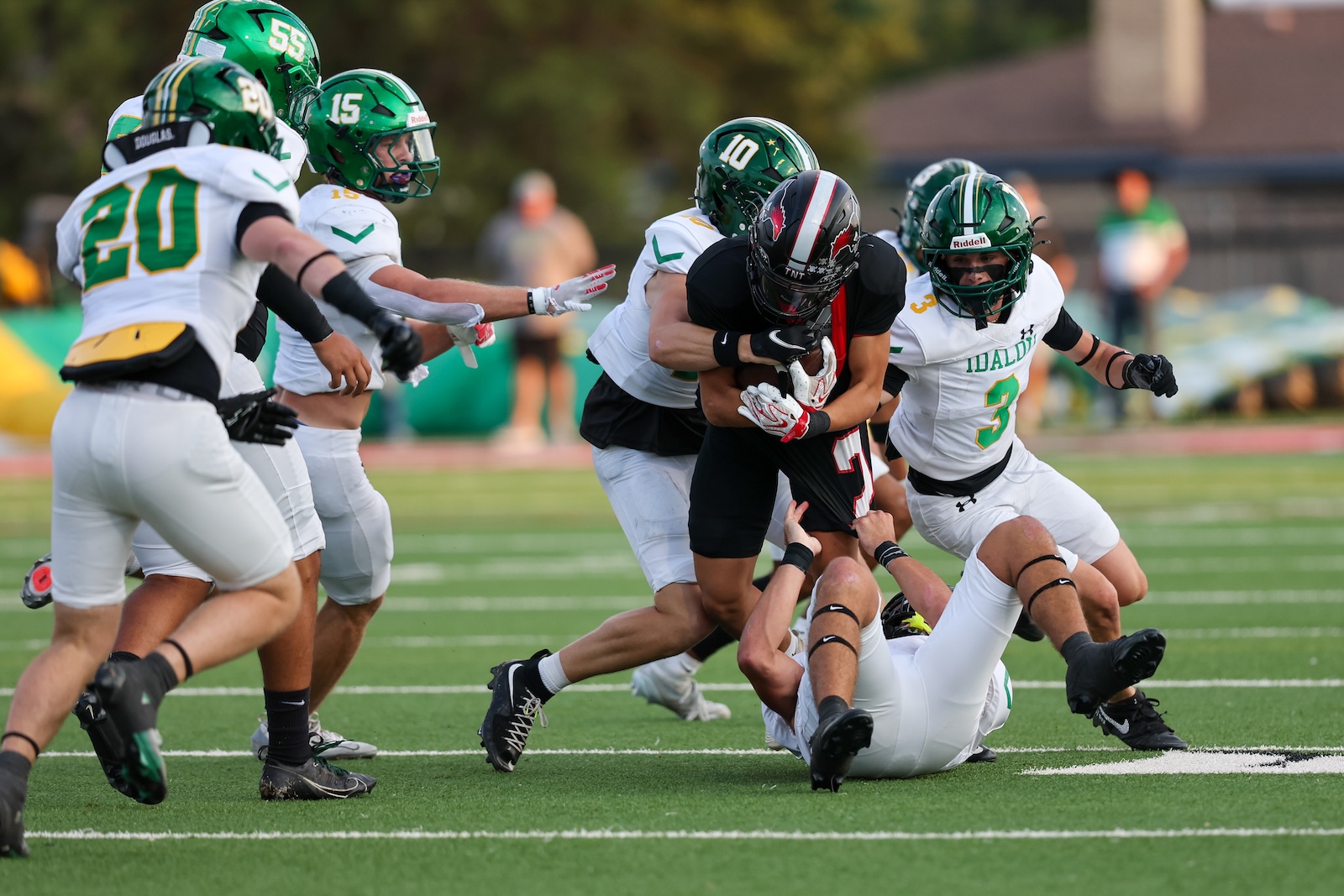 Seabolt Chase-16952 | Press Pass Sports Football action: a ball carrier in black is tackled by multiple defenders in white uniforms with green helmets on a green turf field