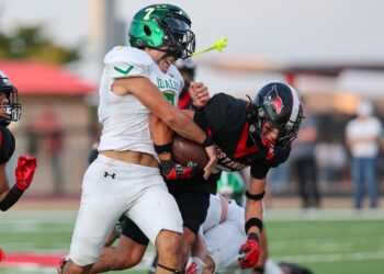 Two football players collide on the field: a ball carrier in a white jersey is tackle‑driven by a defender in black and red.