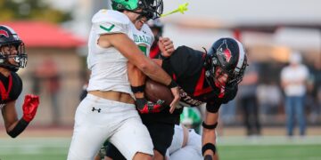 Two football players collide on the field: a ball carrier in a white jersey is tackle‑driven by a defender in black and red.