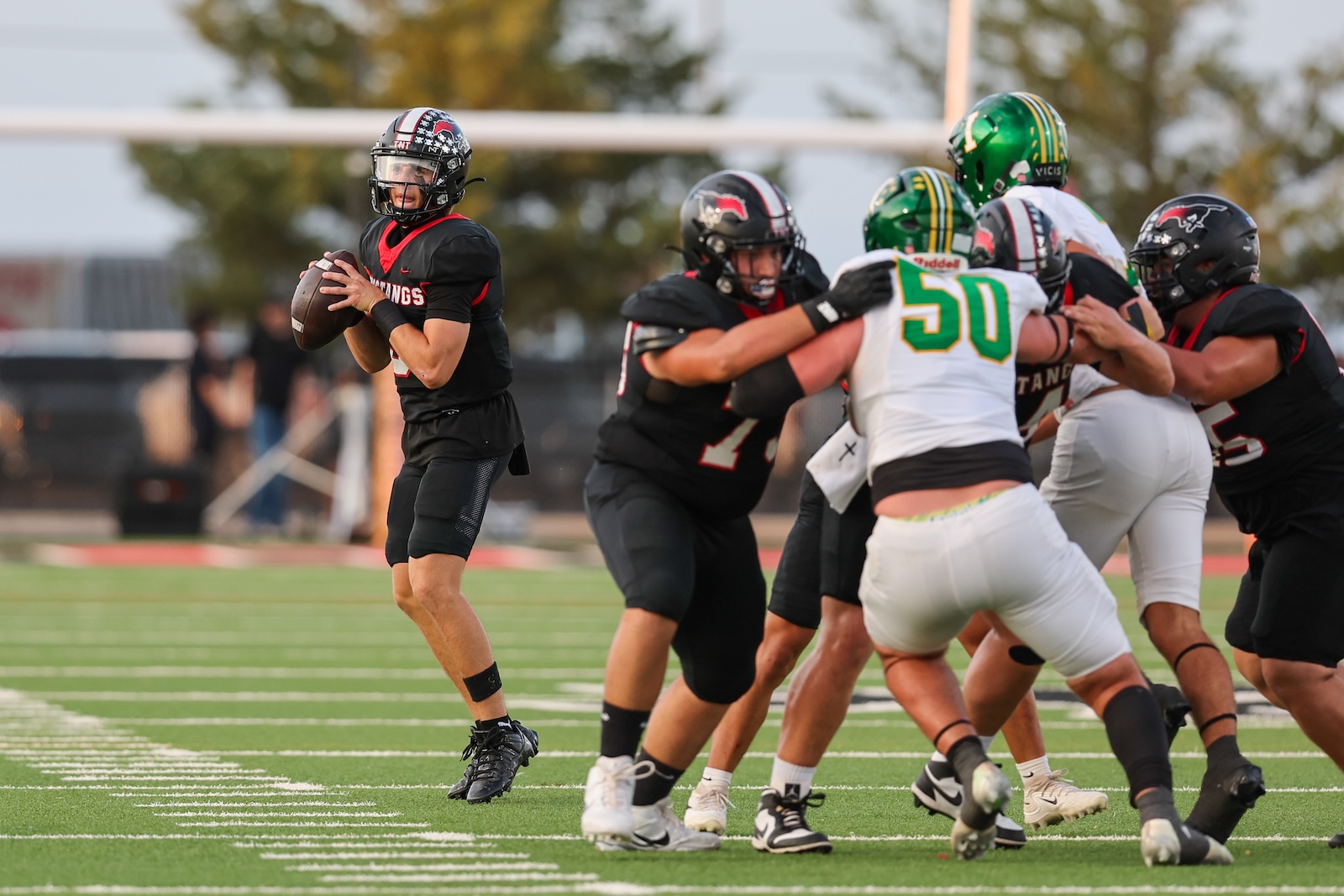 Seabolt Chase-17076 | Press Pass Sports Football quarterback in black uniform winds up to throw, teammates and defenders collide in the background on a green field.