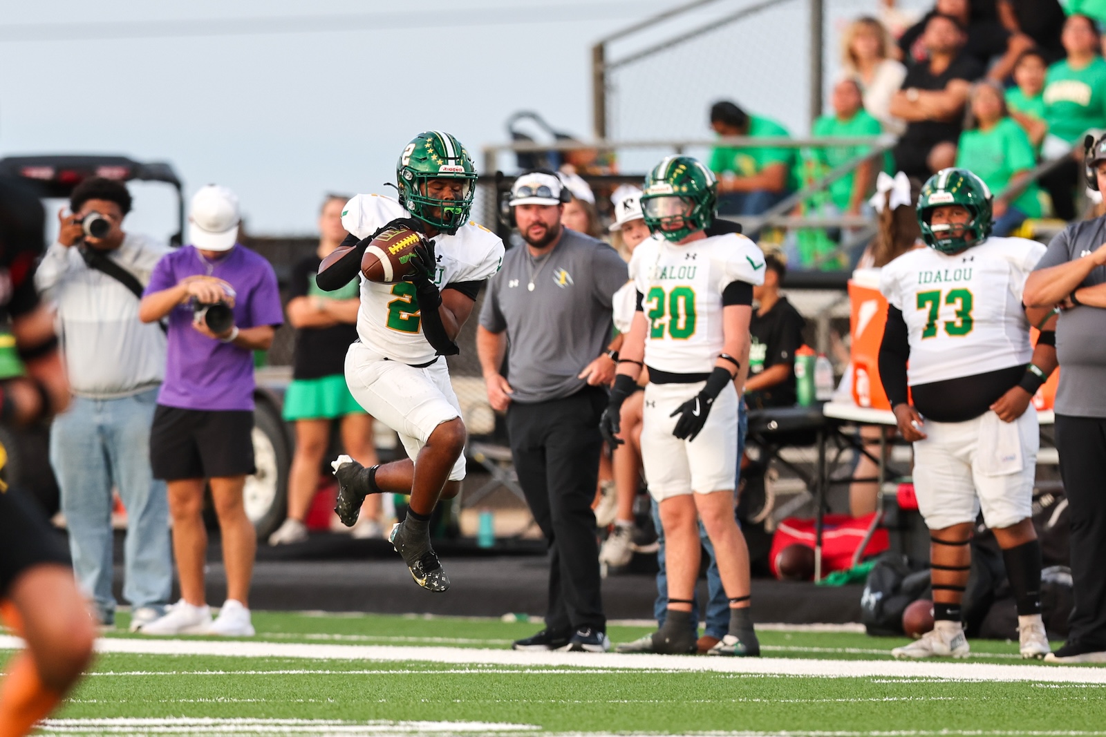 Seabolt Chase-17131 | Press Pass Sports Receiver in white-green uniform leaps to catch a short pass near the sideline as photographers and teammates watch in the background on a football field.