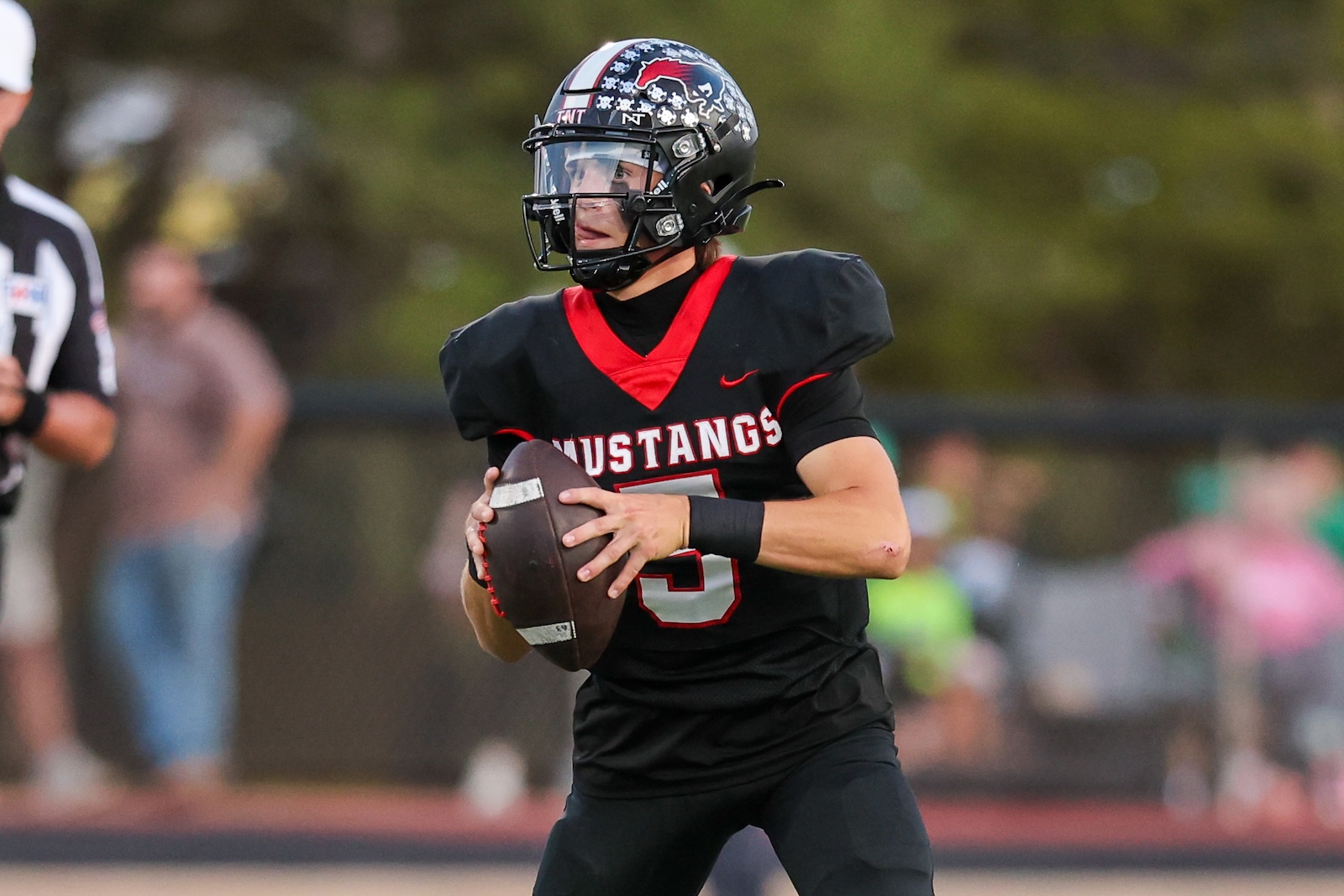 Seabolt Chase-17322 | Press Pass Sports Football player in a black and red Mustang uniform grips a football while running on the field during a game, helmet decorated with team logo