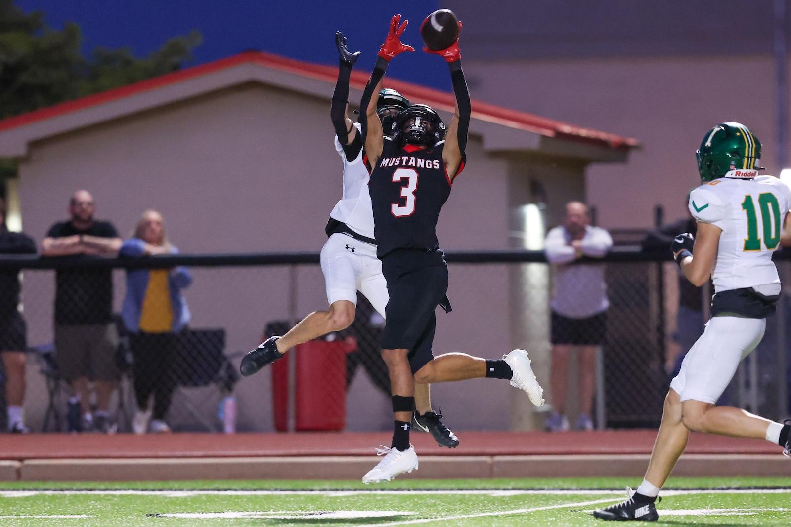 Seabolt Chase-18085 | Press Pass Sports Football players leap for the ball as Mustang #3 reaches up with gloves in a skyward catch attempt on a green turf.