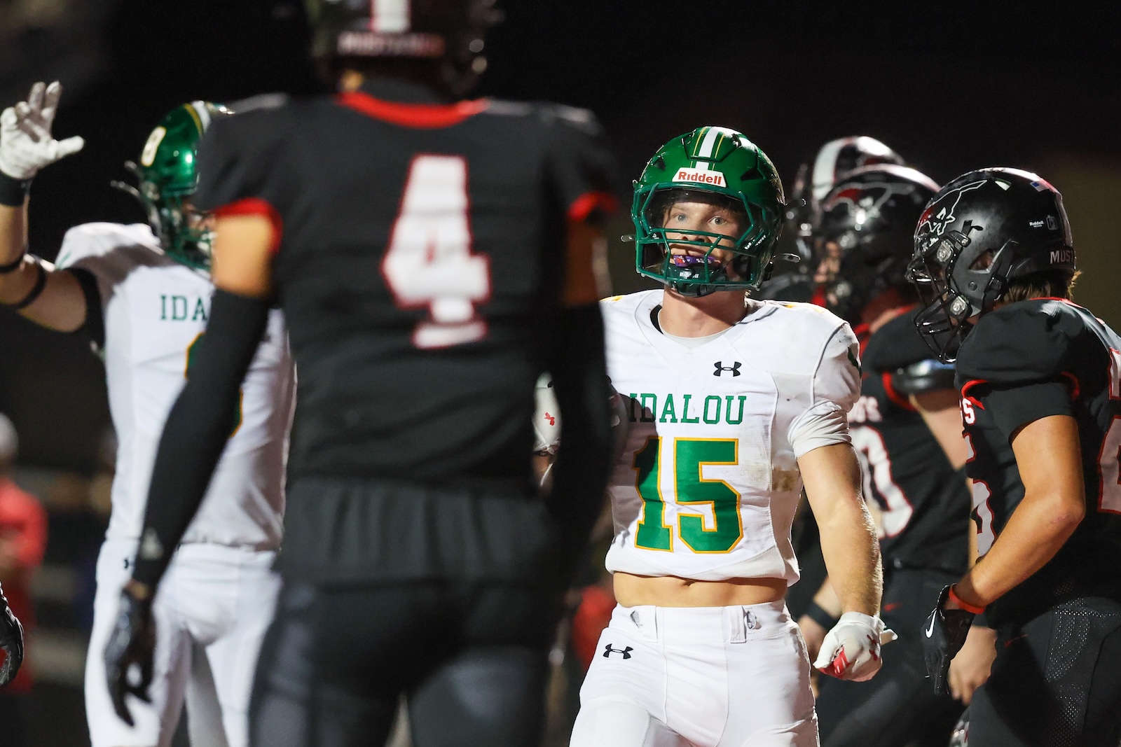 Seabolt Chase-18206 | Press Pass Sports Football players on a nighttime field; center foreground wears white jersey with number 15 and green helmet, facing the camera.