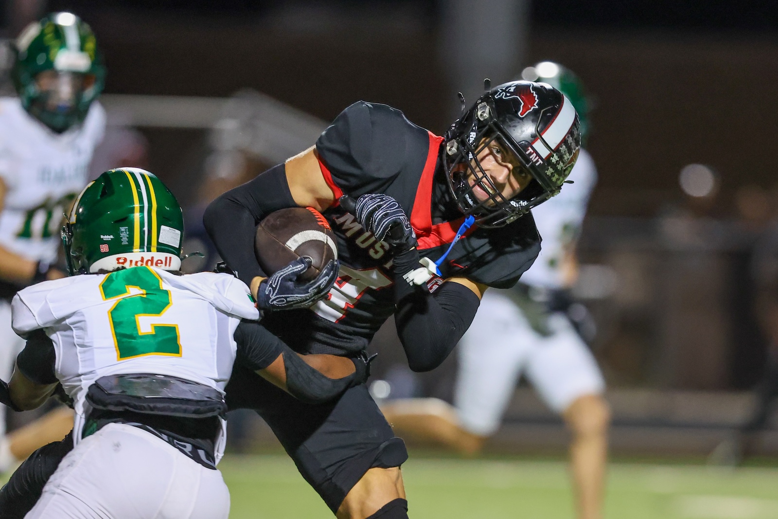 Seabolt Chase-18589 | Press Pass Sports Football player in black jersey with red trim carries the ball while being tackled by a defender in a white uniform with green helmet at night.
