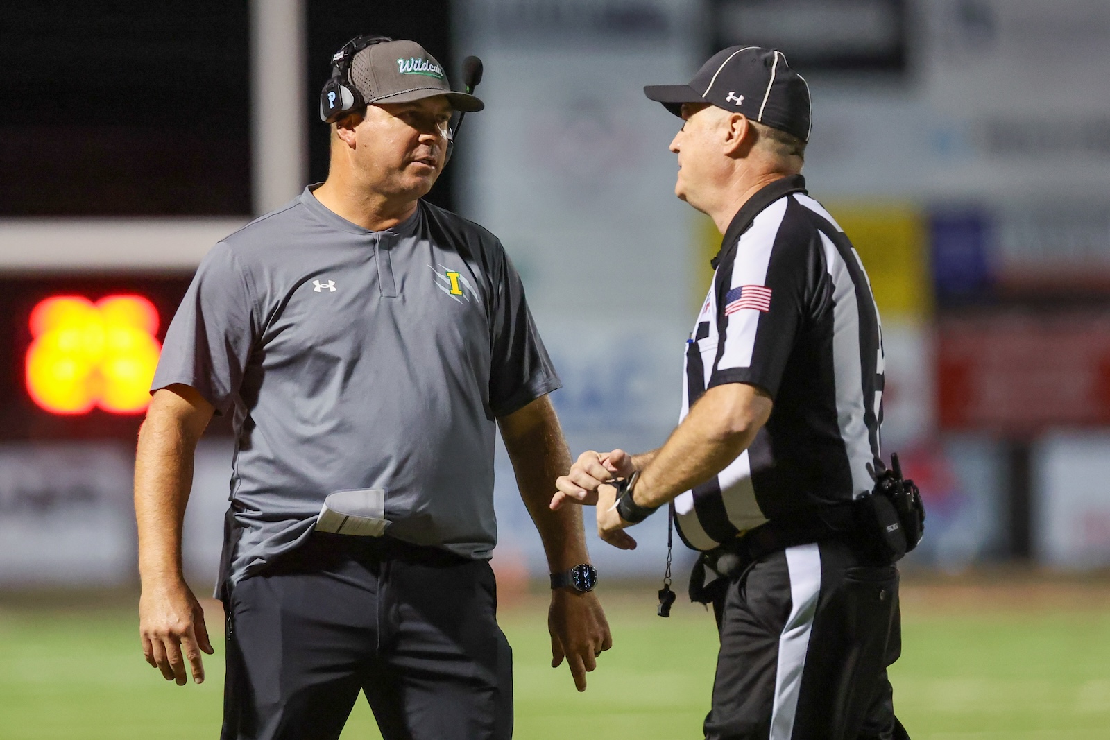 Seabolt Chase-18737 | Press Pass Sports Two men on a football field shake hands, one in a gray polo with a headset and the other in a black-and-white striped referee uniform at night.