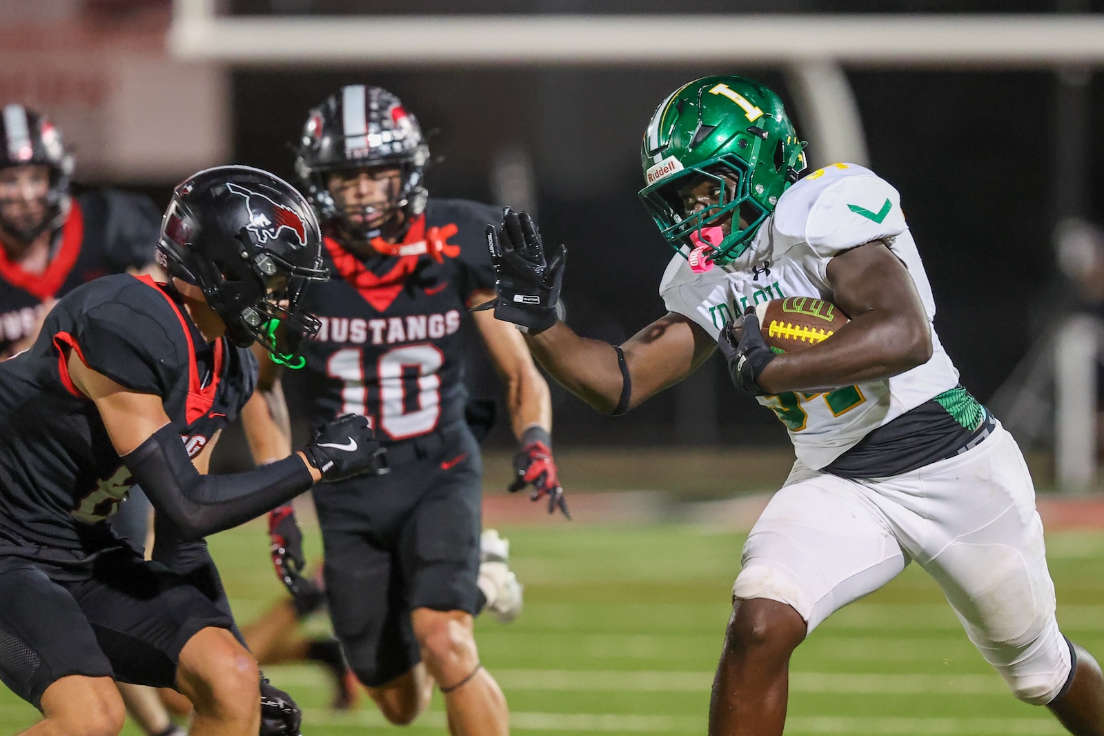 34N | Press Pass Sports Football player in white and green with the ball runs forward while defenders in black close in. Night game on a grassy field.