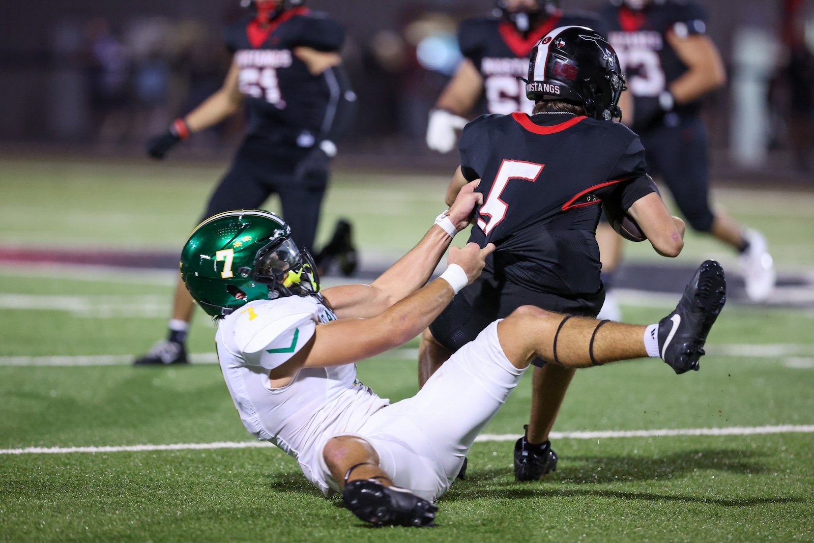 Seabolt Chase-19083 | Press Pass Sports Tackle scene: defender in white and green brings down the ball carrier in a black jersey (#5) on a football field at night.