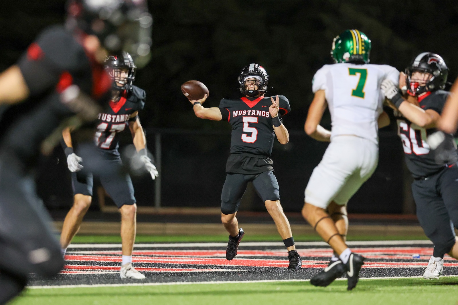 Seabolt Chase-19218 | Press Pass Sports Quarterback in black Mustang jersey number 5 throws a pass while teammates and defenders converge on the field at night.
