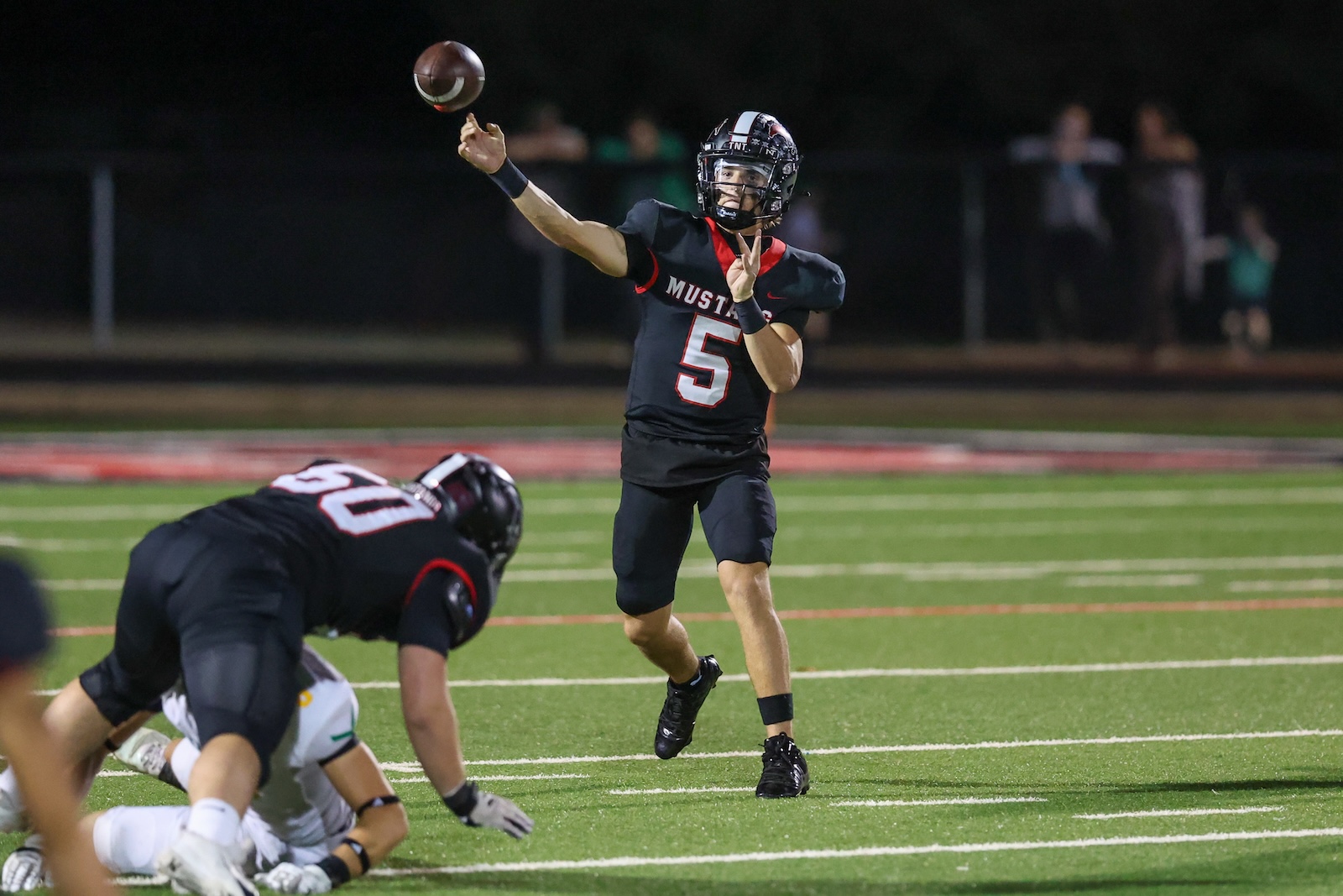 Seabolt Chase-19315 | Press Pass Sports Quarterback in a black uniform (#5) throws a pass during a night football game on a green turf field.