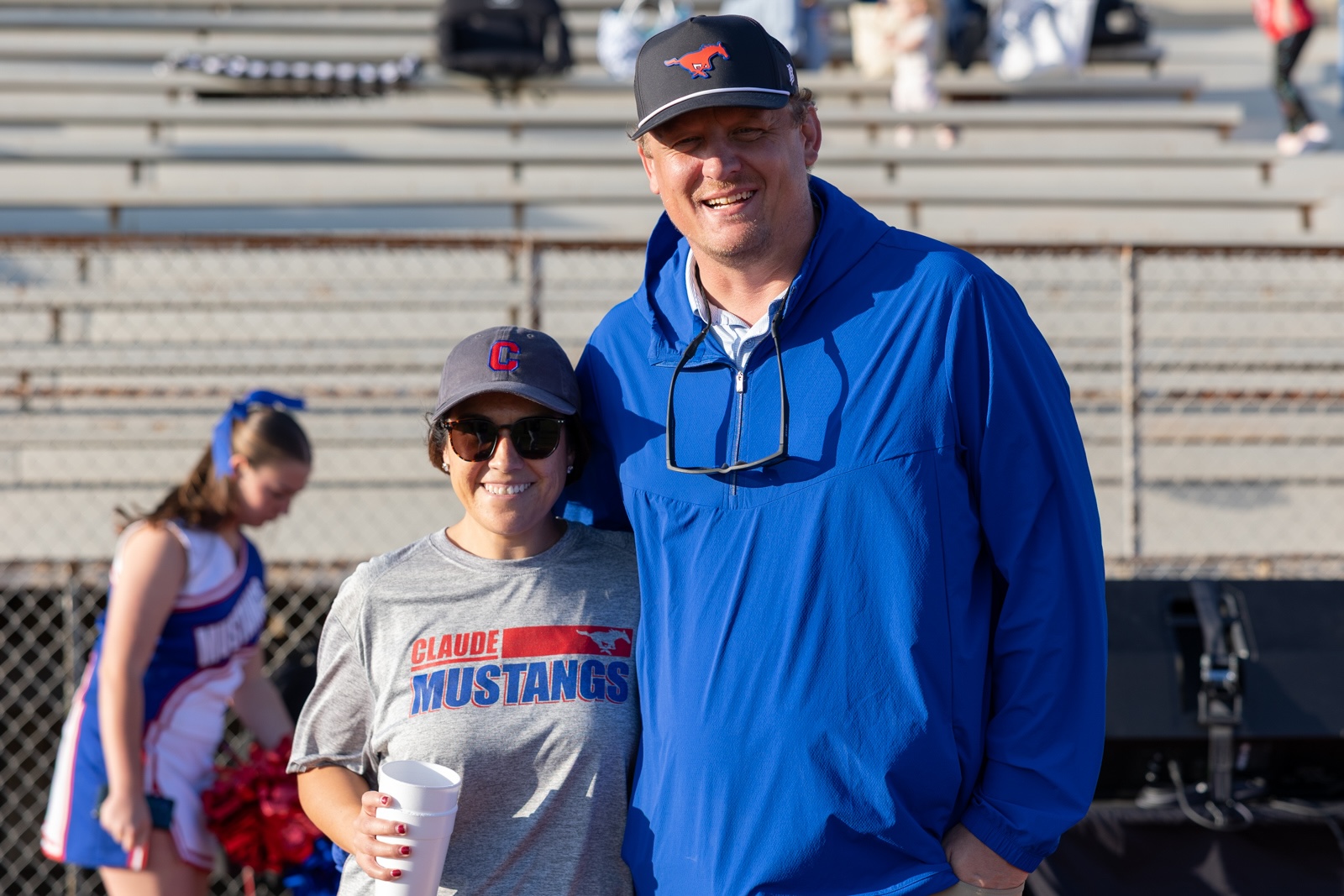 Liberty_15-4 | Press Pass Sports Two people posing for a photo at a stadium: a woman in a gray Claude Mustangs shirt and sunglasses next to a man in a bright blue jacket, with bleachers behind them and a cheerleader in the background.
