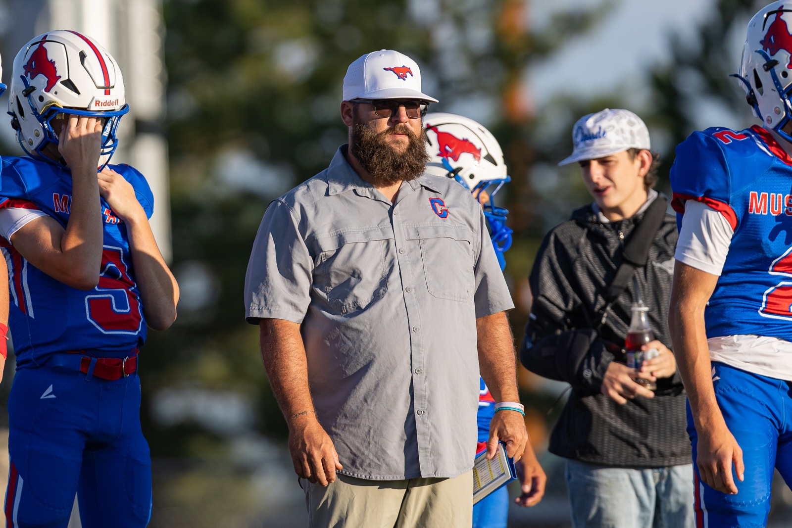 Liberty_15-5 | Press Pass Sports Bearded coach in a gray shirt and white cap with a red logo stands among football players in blue uniforms on the field.