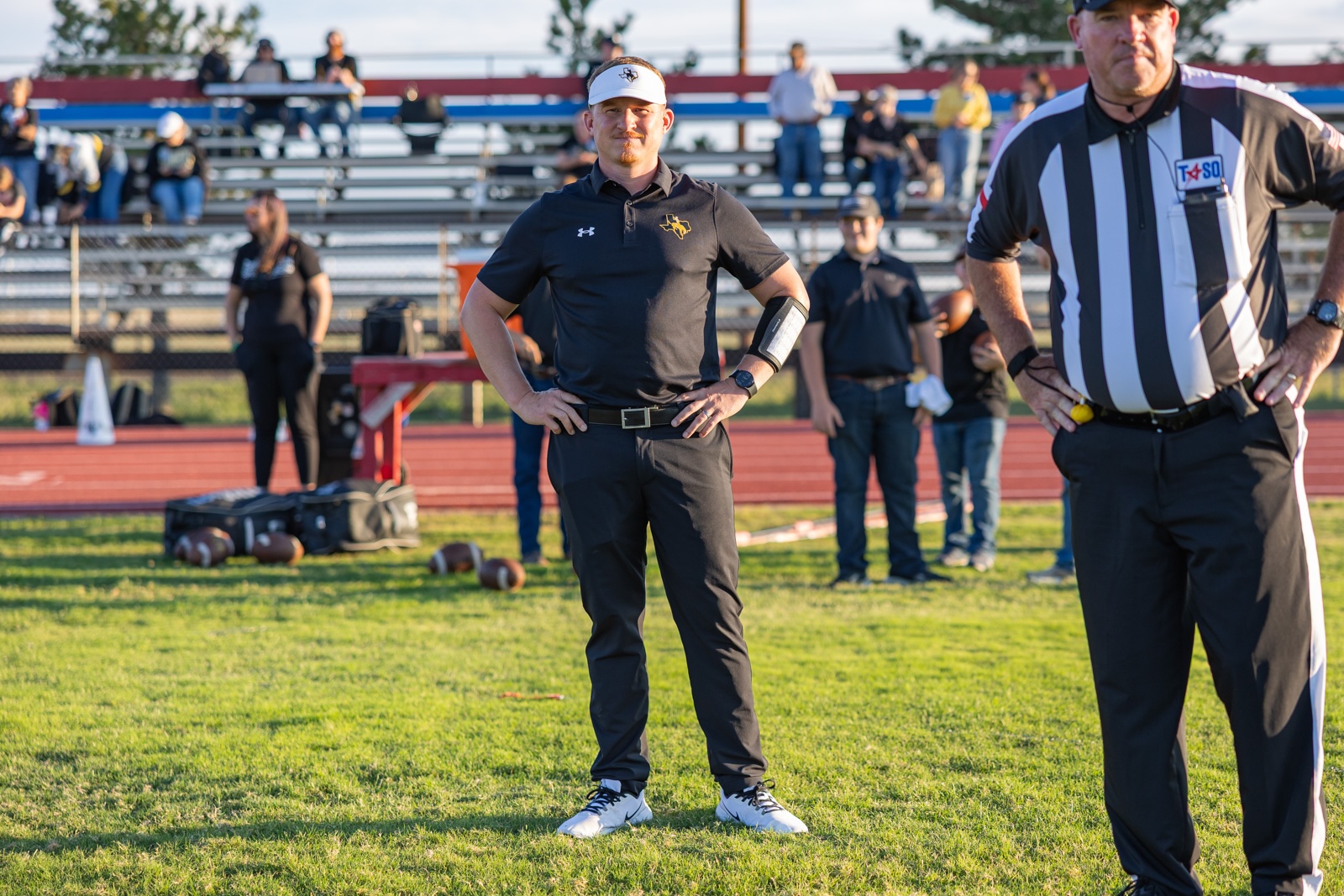 Liberty_15-6 | Press Pass Sports Man in black polo and white visor stands with hands on hips on a football field, referee in striped shirt to the right, bleachers and people in the background.