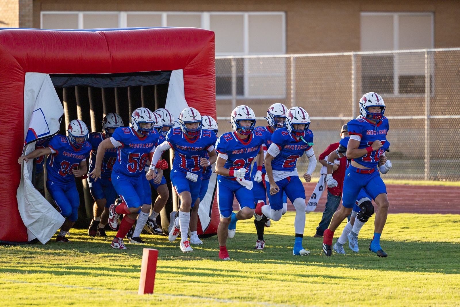 Liberty_15-7 | Press Pass Sports High school football players in blue uniforms sprint from a red inflatable tunnel onto a grassy field.
