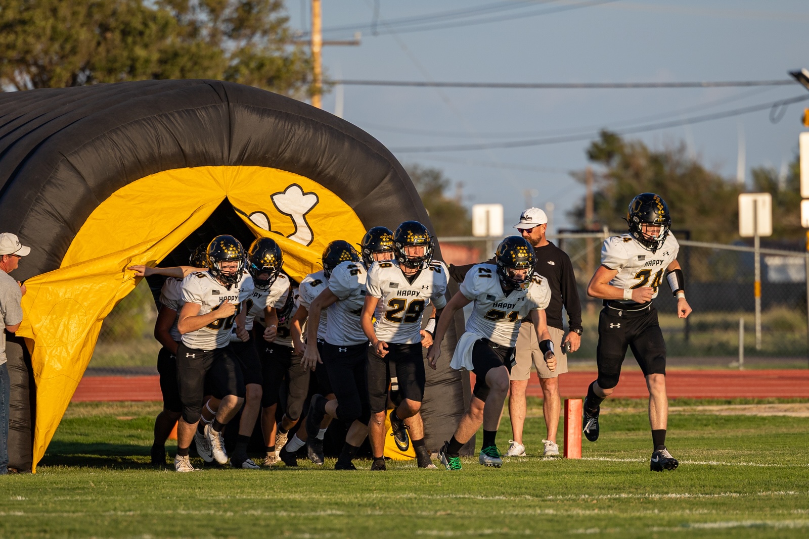 Liberty_15-8 | Press Pass Sports Football players in white uniforms sprint onto the field from a yellow and black inflatable tunnel behind them.