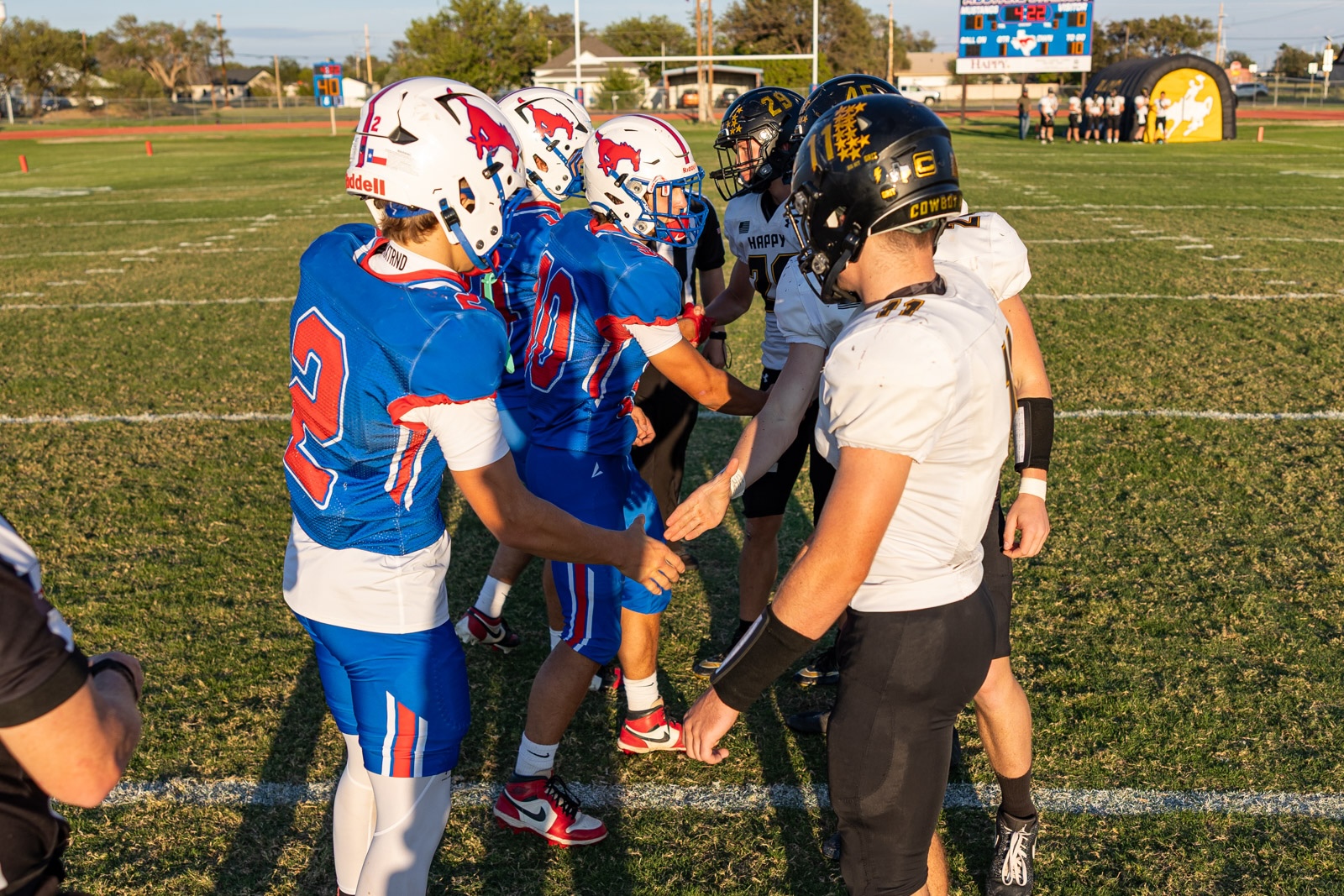 Liberty_15-9 | Press Pass Sports Football players in blue and white uniforms shake hands in a pre-game line on a sunny grassy field with a scoreboard in the background.
