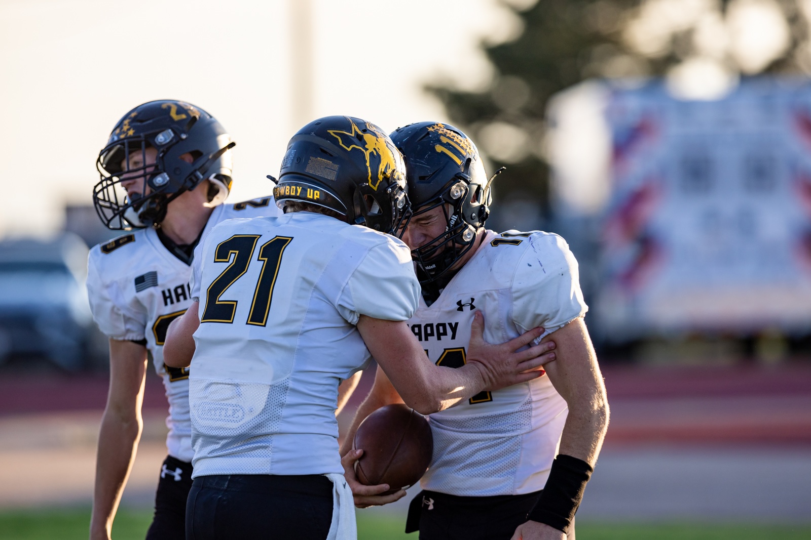 Liberty_15-12 | Press Pass Sports Three football players in white uniforms hustle together on the field, one holding the ball as they celebrate or plan a play edge to edge hug.