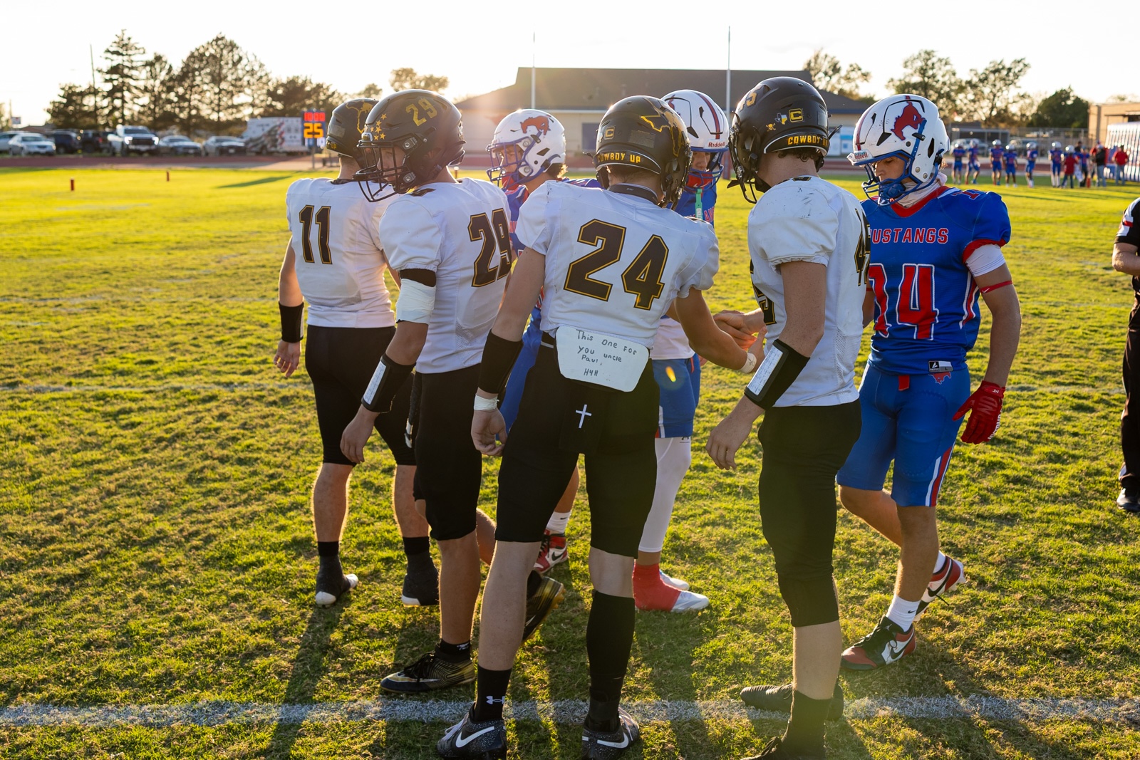 Liberty_15-14 | Press Pass Sports Football players in white uniforms form a pregame huddle on a sunny field, numbers 11, 24 visible on backs.