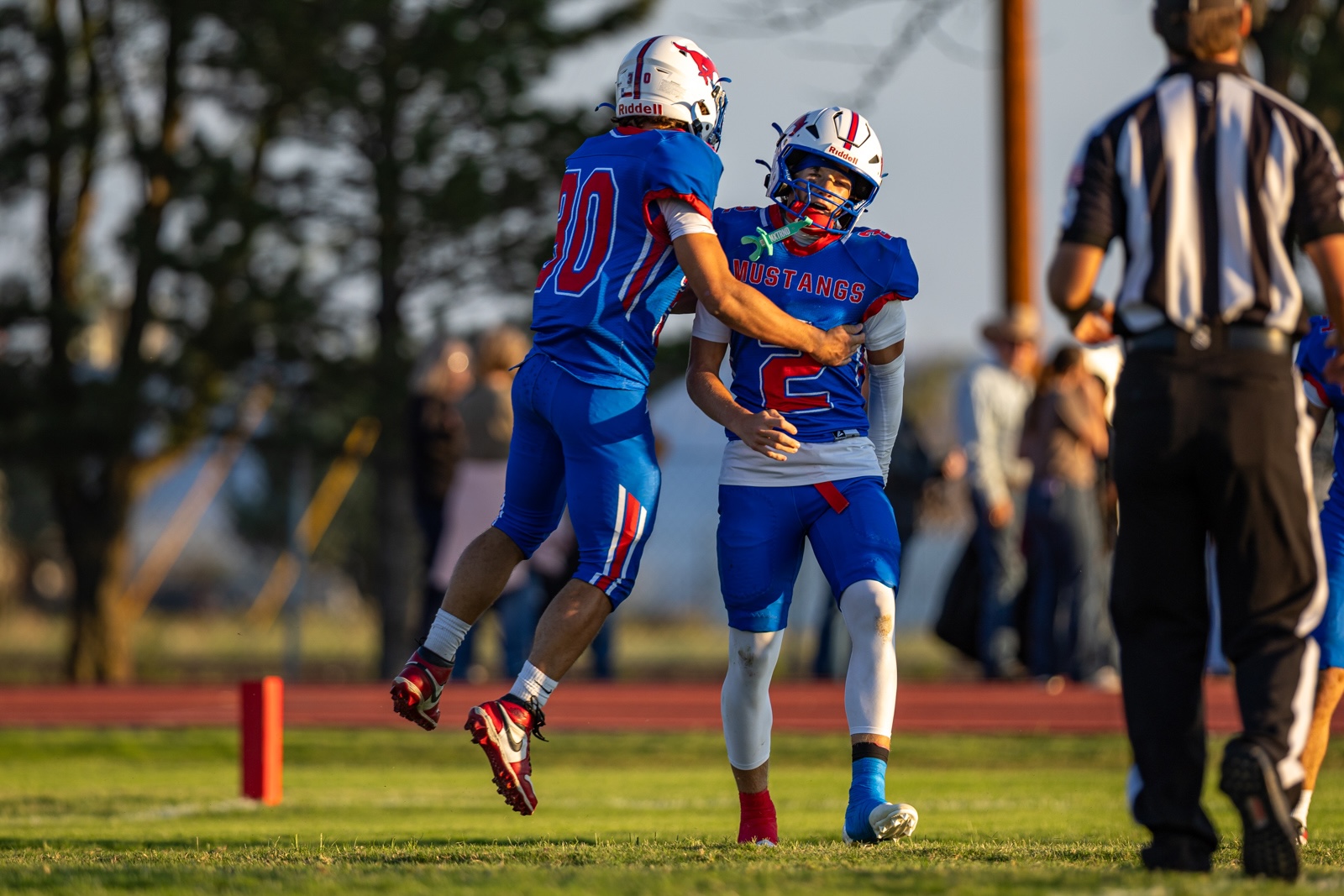 Liberty_15-16 | Press Pass Sports Two football players in blue Mustangs uniforms celebrate on a grass field, one leaping toward the other in mid‑celebration.