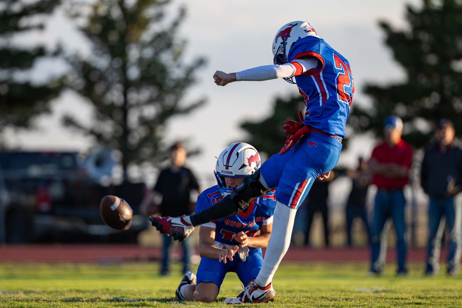 Liberty_15-17 | Press Pass Sports Two football players in blue uniforms collide on a grassy field as one leaps in the air while another reaches for the ball nearby; spectators blur in the background.