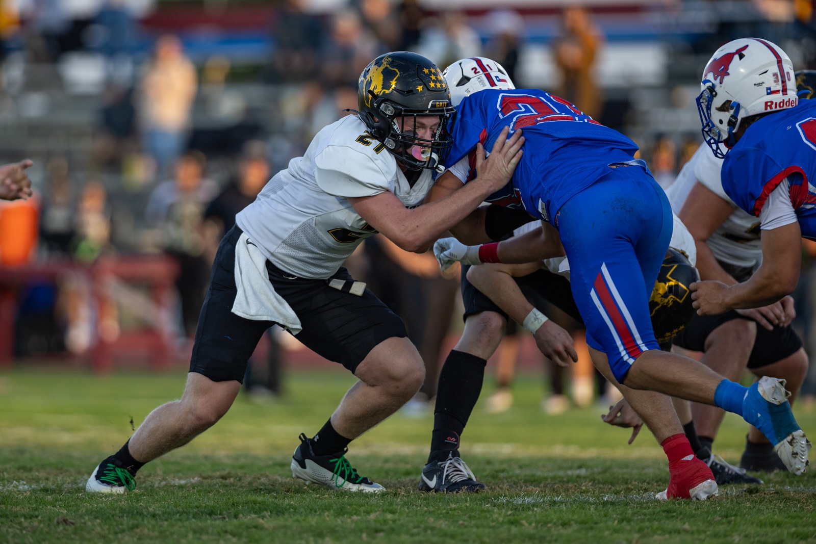 Liberty_15-18 | Press Pass Sports Two football players collide at the line of scrimmage, one in white with a dark helmet and the other in blue, on a grassy field