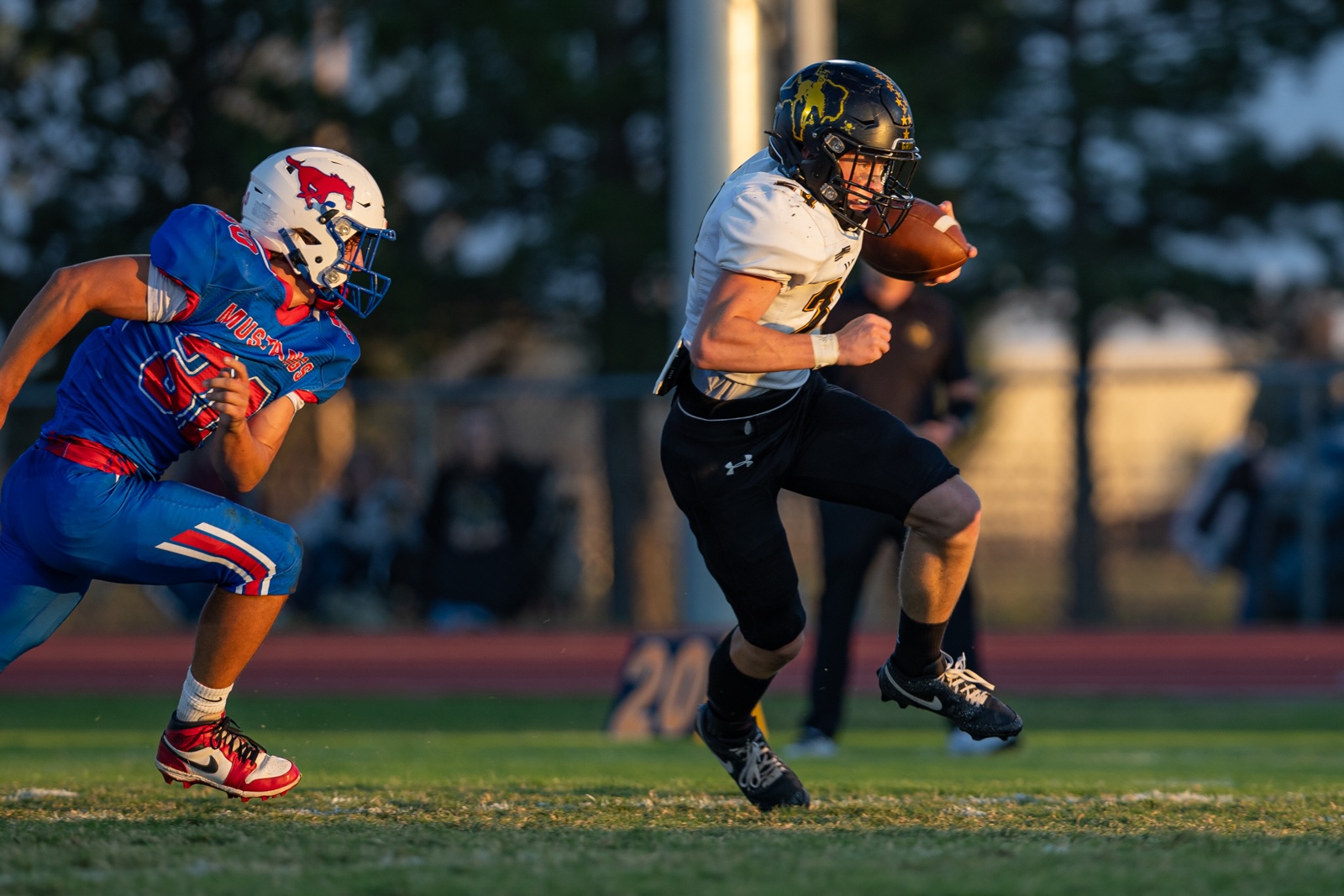 Liberty_15-19 | Press Pass Sports Football players sprinting; a blue-uniform defender chases a white-jersey ball carrier with the ball tucked underarm on a sunny field.