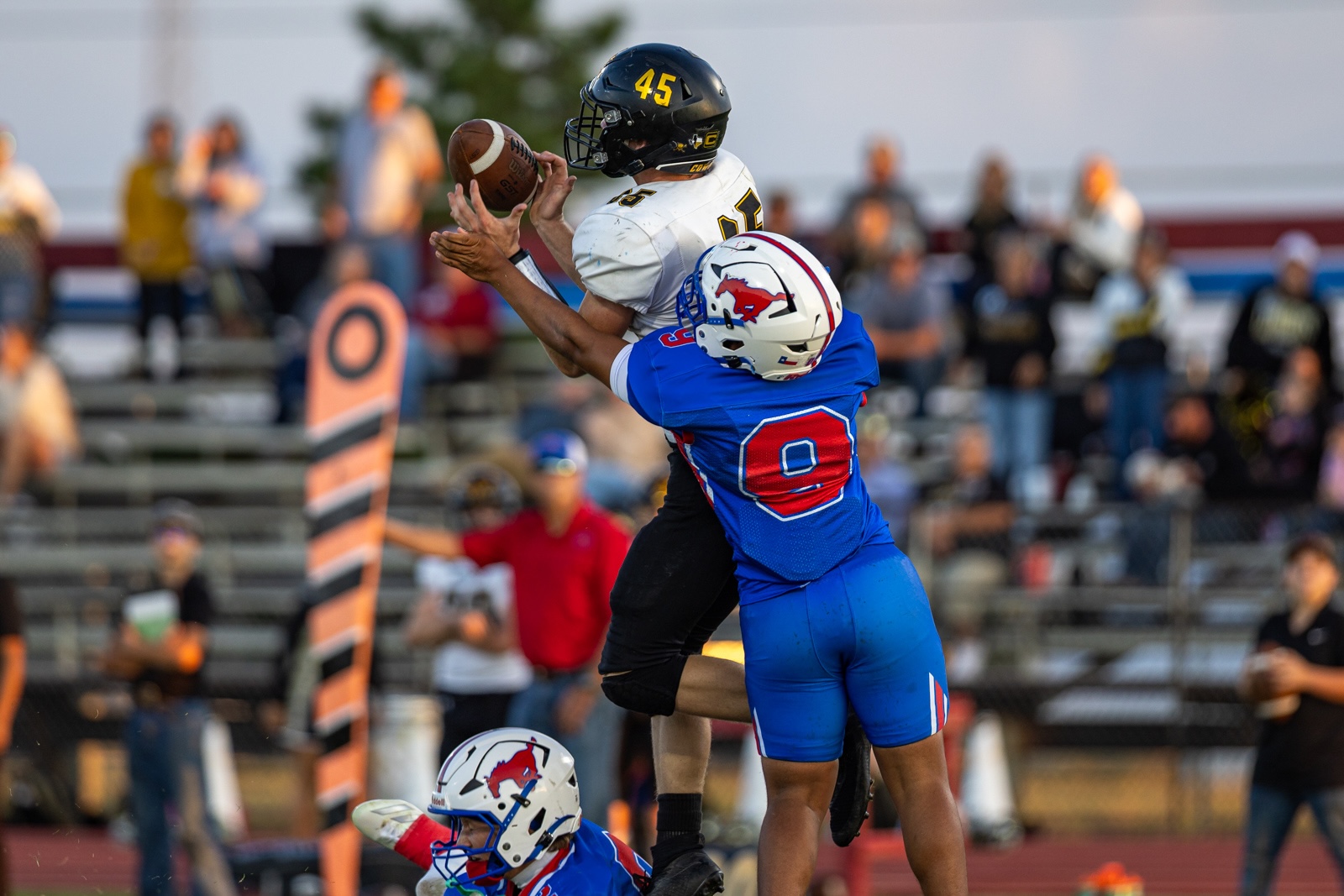 Liberty_15-20 | Press Pass Sports Two football players leap for the ball during a game: the white-jersey player (No. 45) grapples with a blue-jersey defender (No. 9) as they contest the pass.