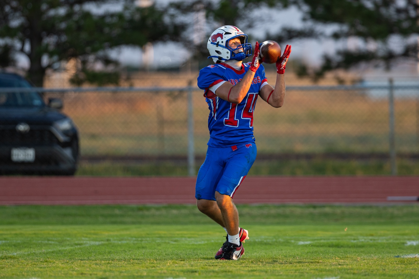 Liberty_15-21 | Press Pass Sports American football player in blue uniform catching a pass on a grassy field, wearing a white helmet with a red logo and red gloves (number 14).